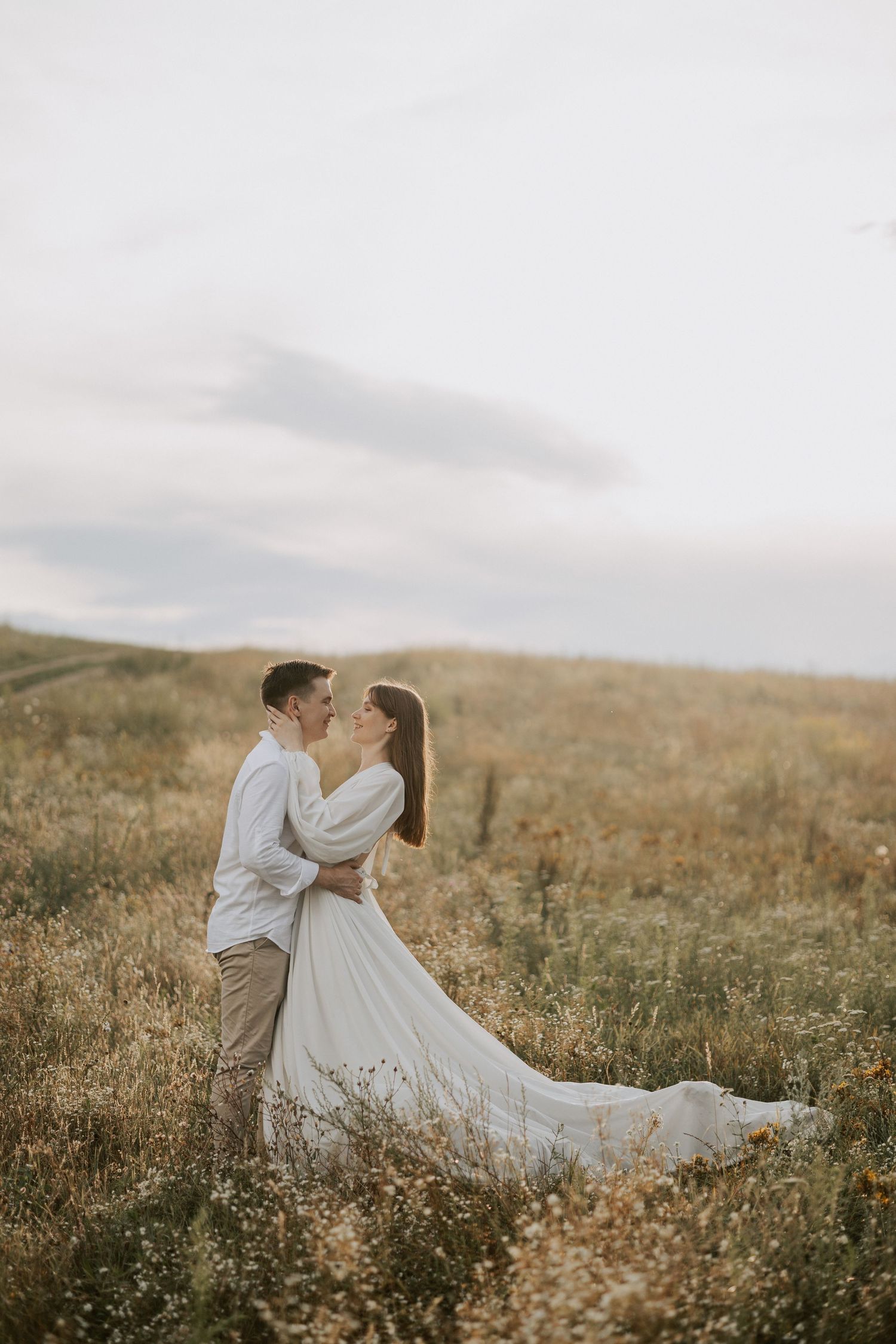 Couple in white clothing share an intimate moment on a hilltop during golden hour.