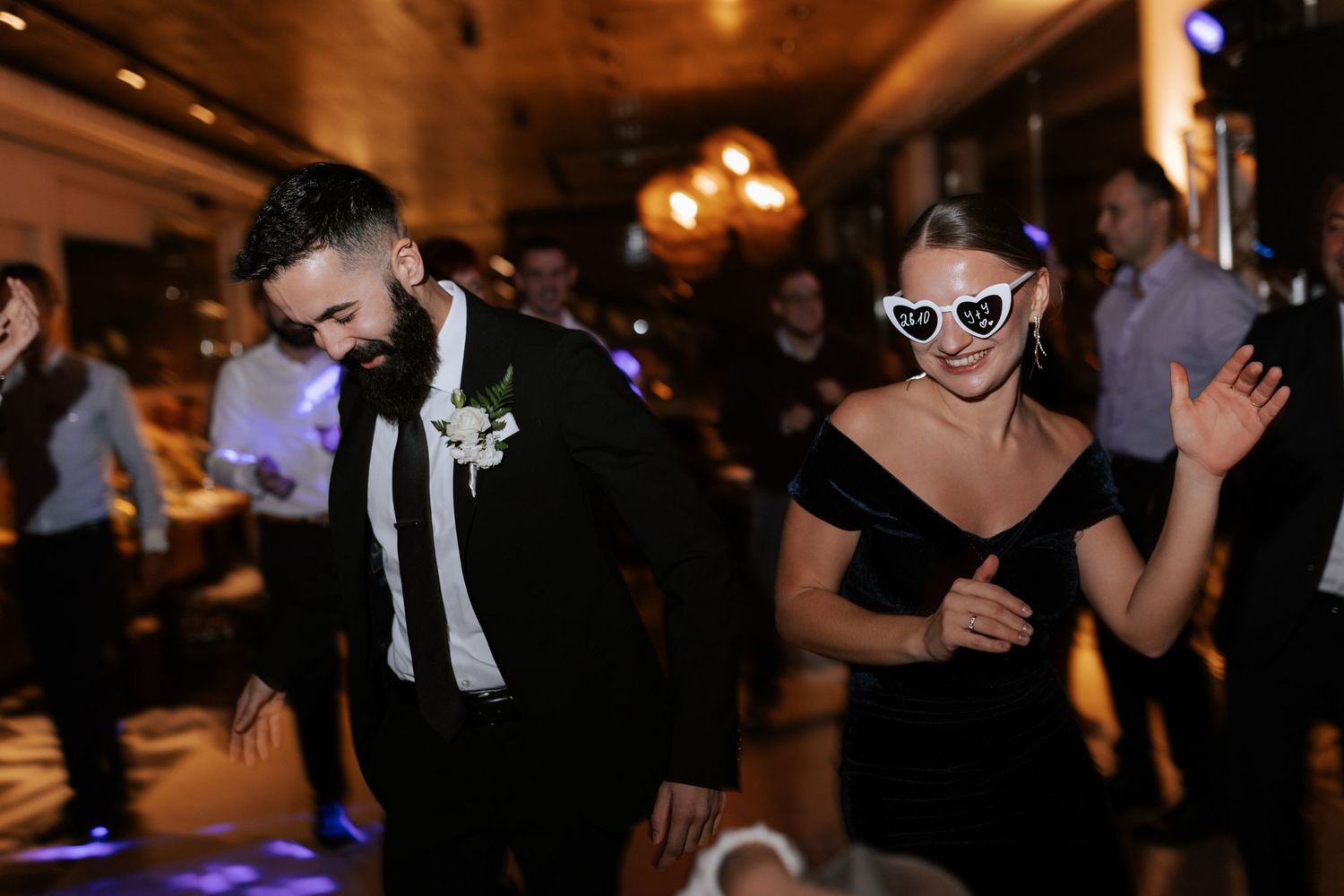 Wedding guests dancing in formal attire with heart sunglasses under moody reception lighting.