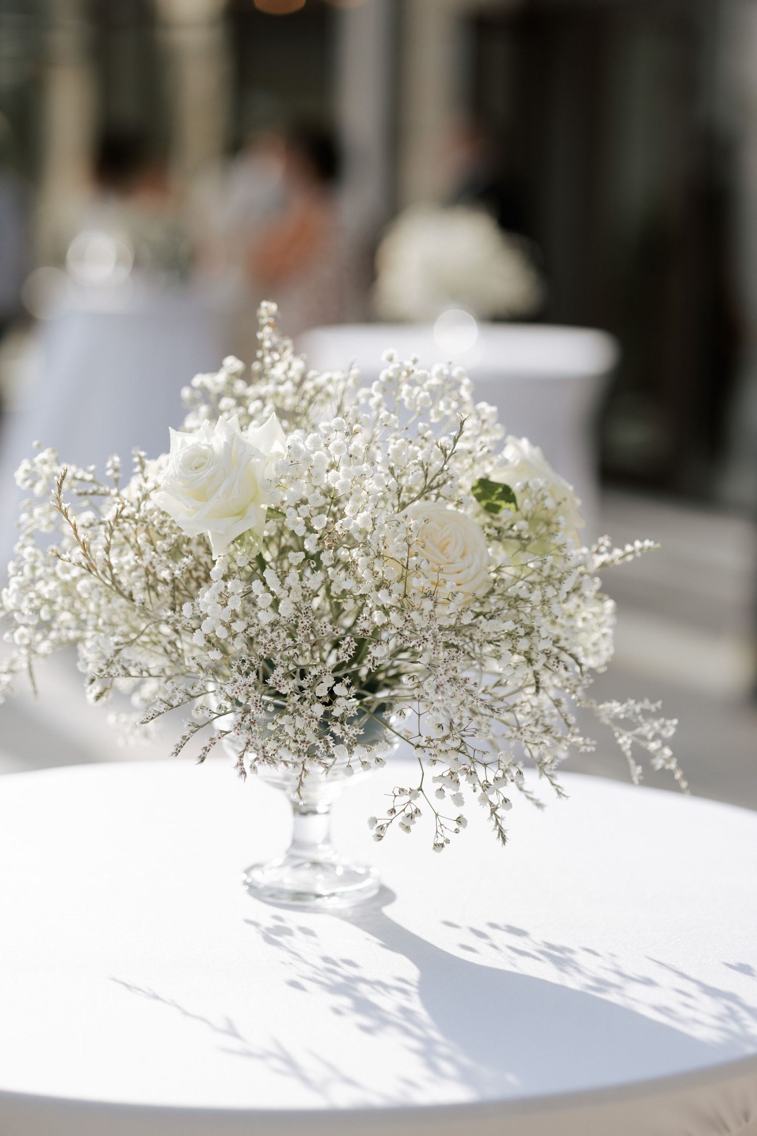 Elegant white floral centerpiece with baby's breath in a glass pedestal vase on a bright white table.