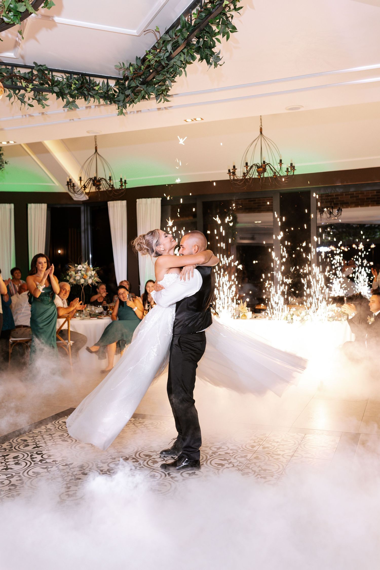 Couple shares their first dance on a misty dance floor with sparklers and green lighting at an elegant indoor wedding reception.