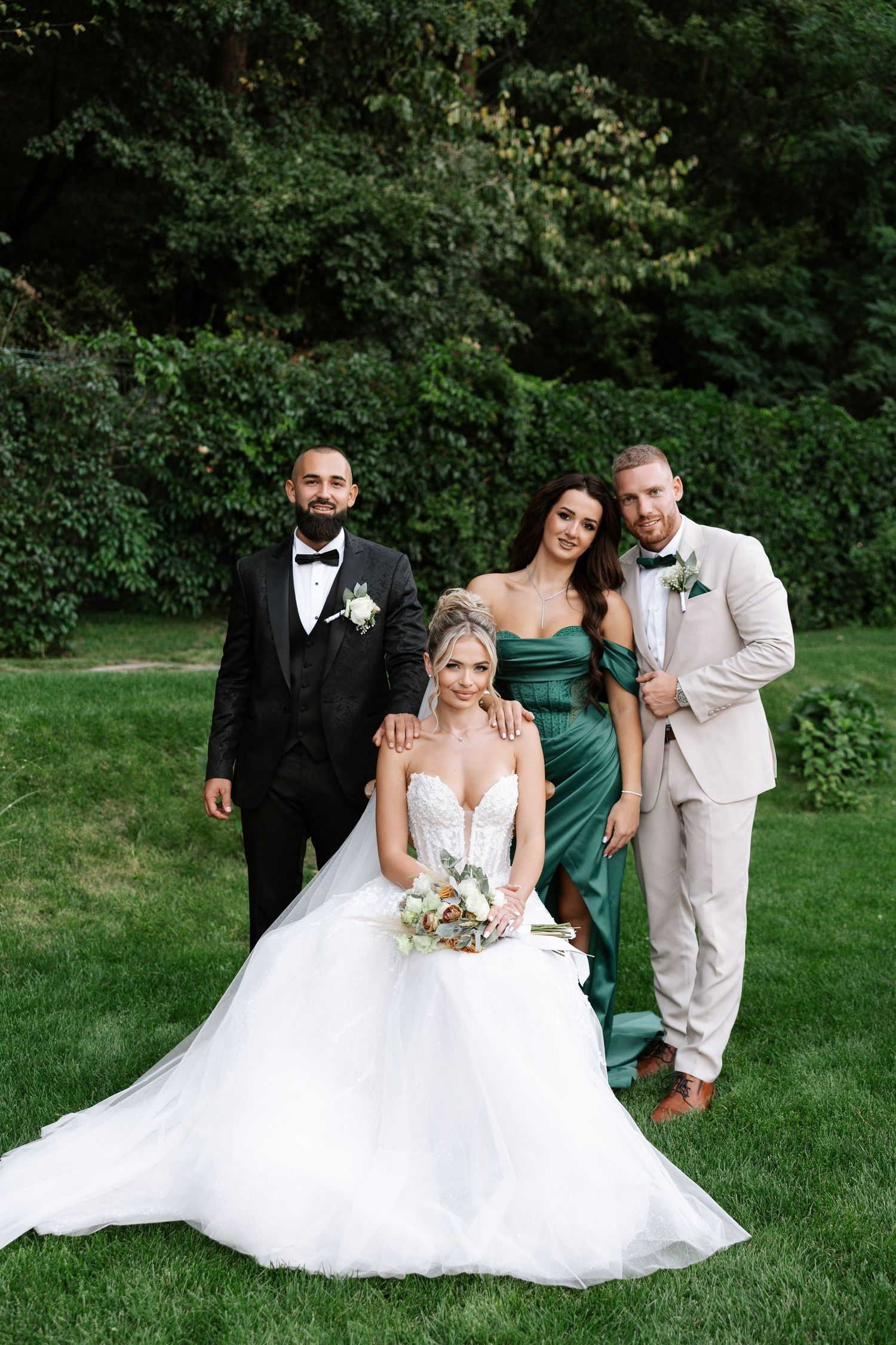 Wedding party poses playfully on lush green lawn with bride in flowing white gown and attendants in formal attire.
