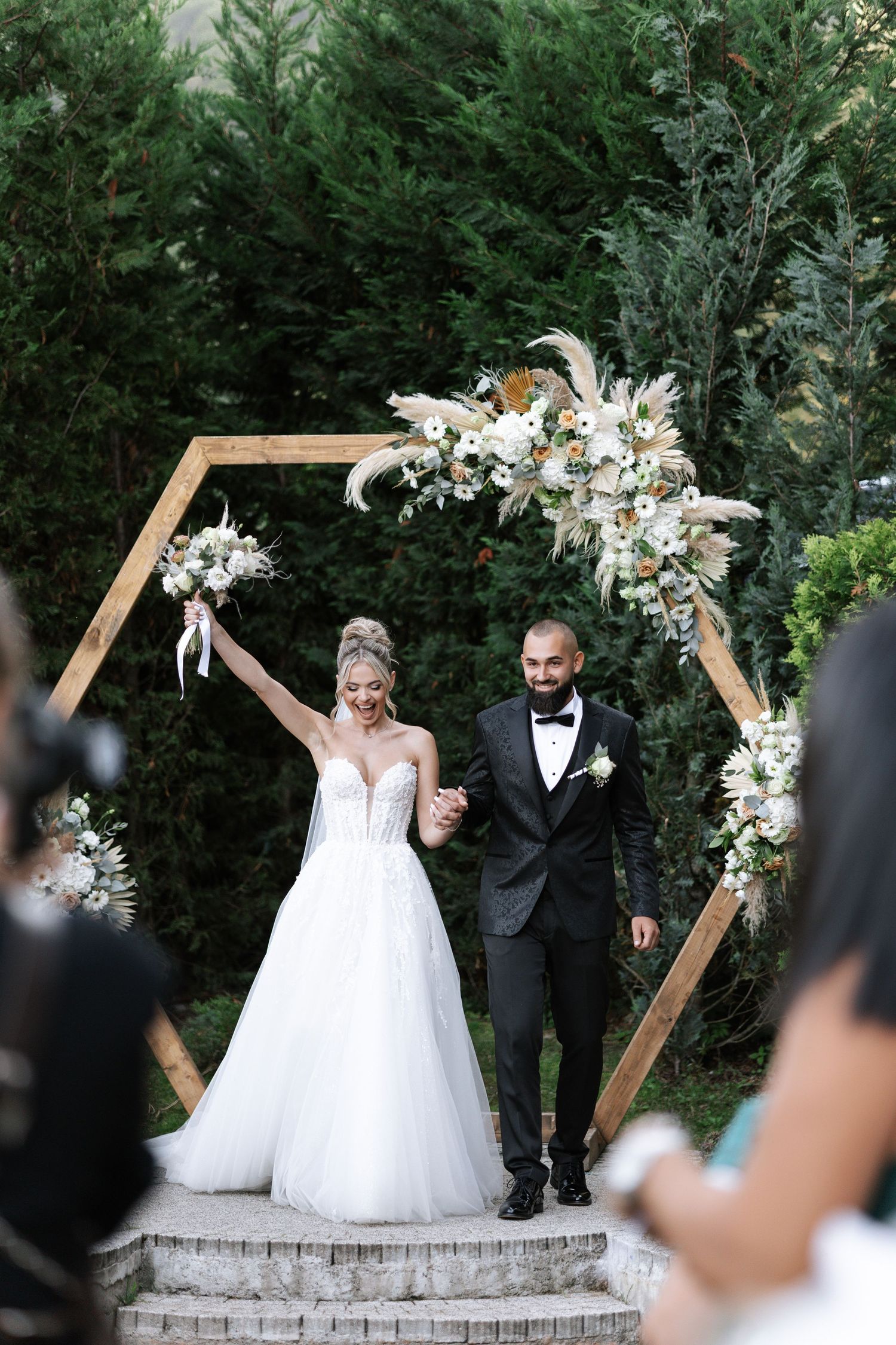 Elegant outdoor wedding ceremony with geometric wooden arch decorated with white lilies and greenery.