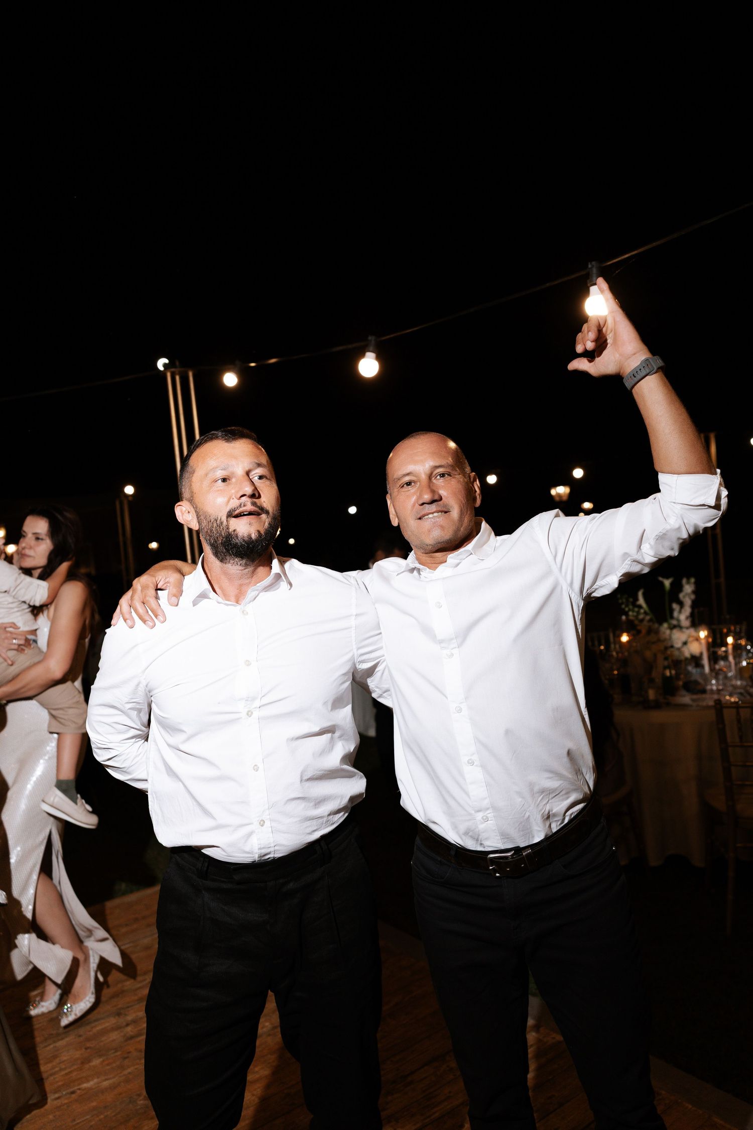 People in white shirts and black pants dance and celebrate at a nighttime outdoor event under string lights.