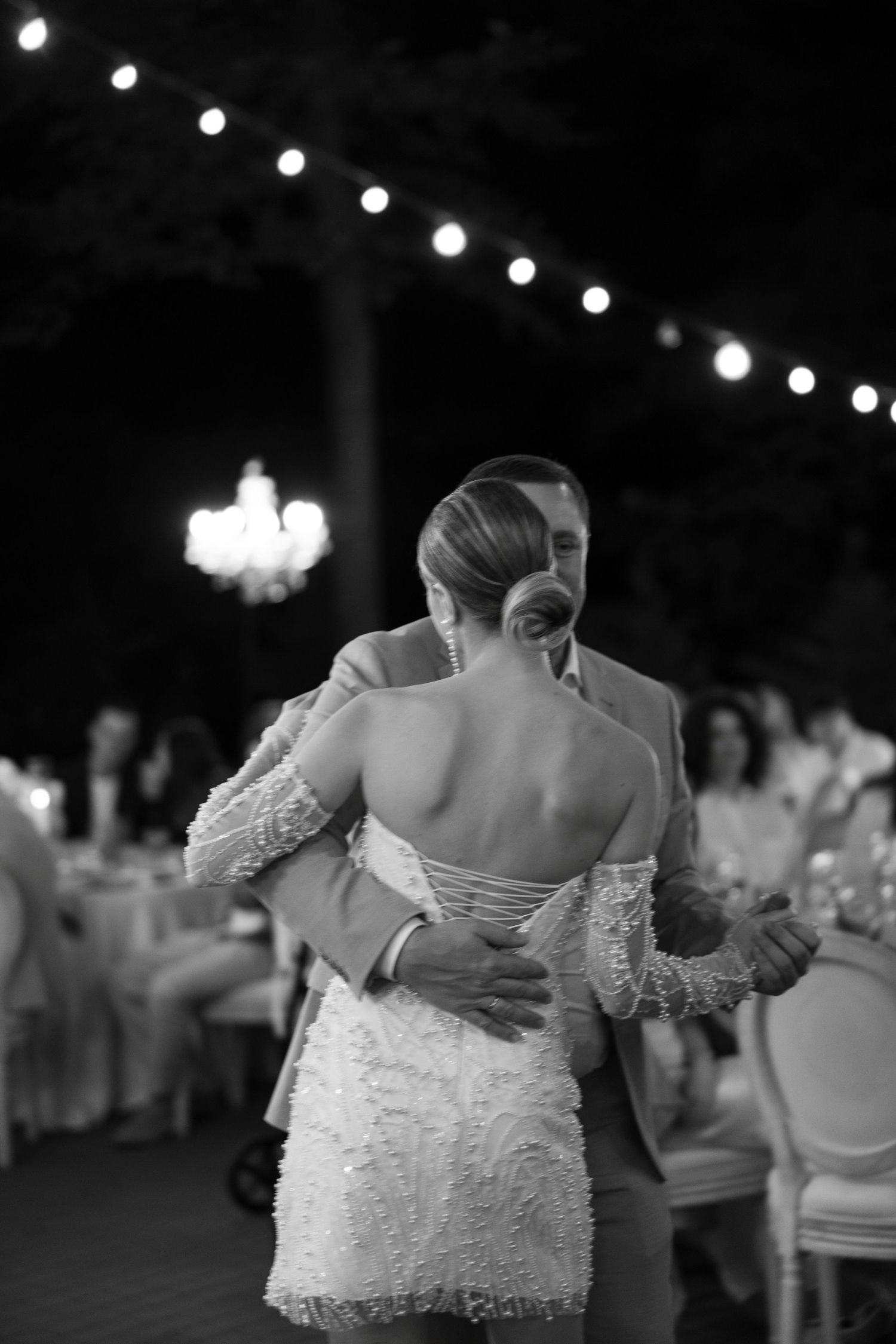 A black and white photo captures an intimate dance moment under string lights at a wedding.