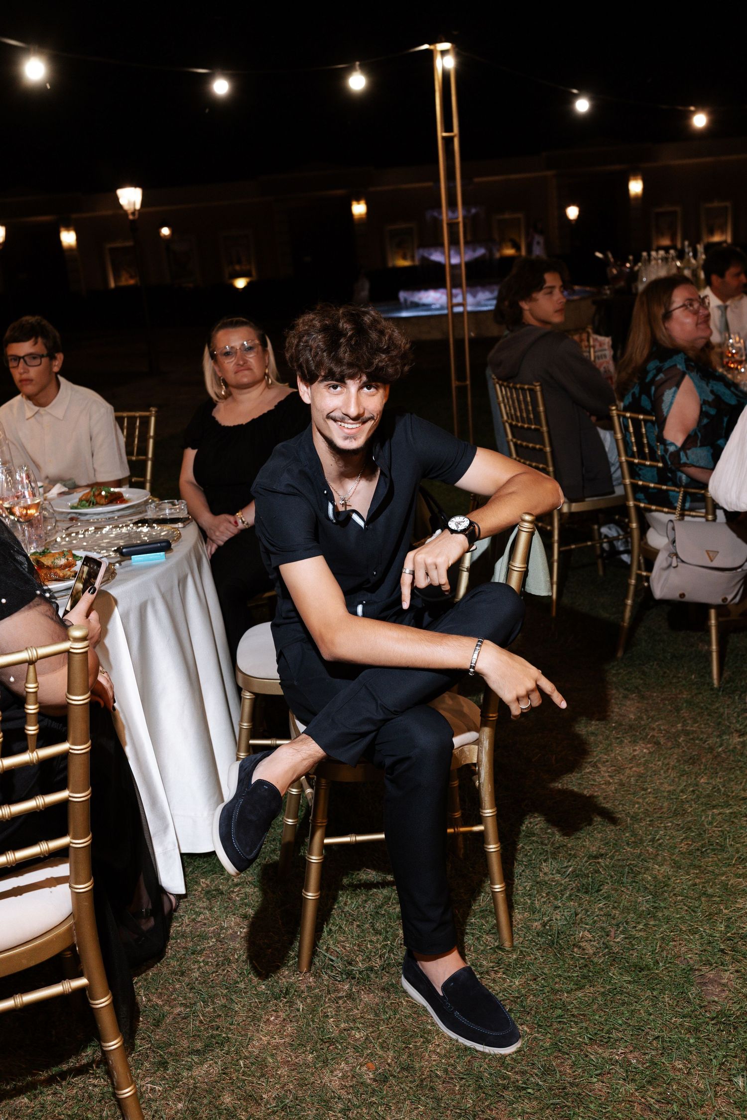 Evening outdoor event with string lights overhead as guests sit at wooden chairs on grass at night.