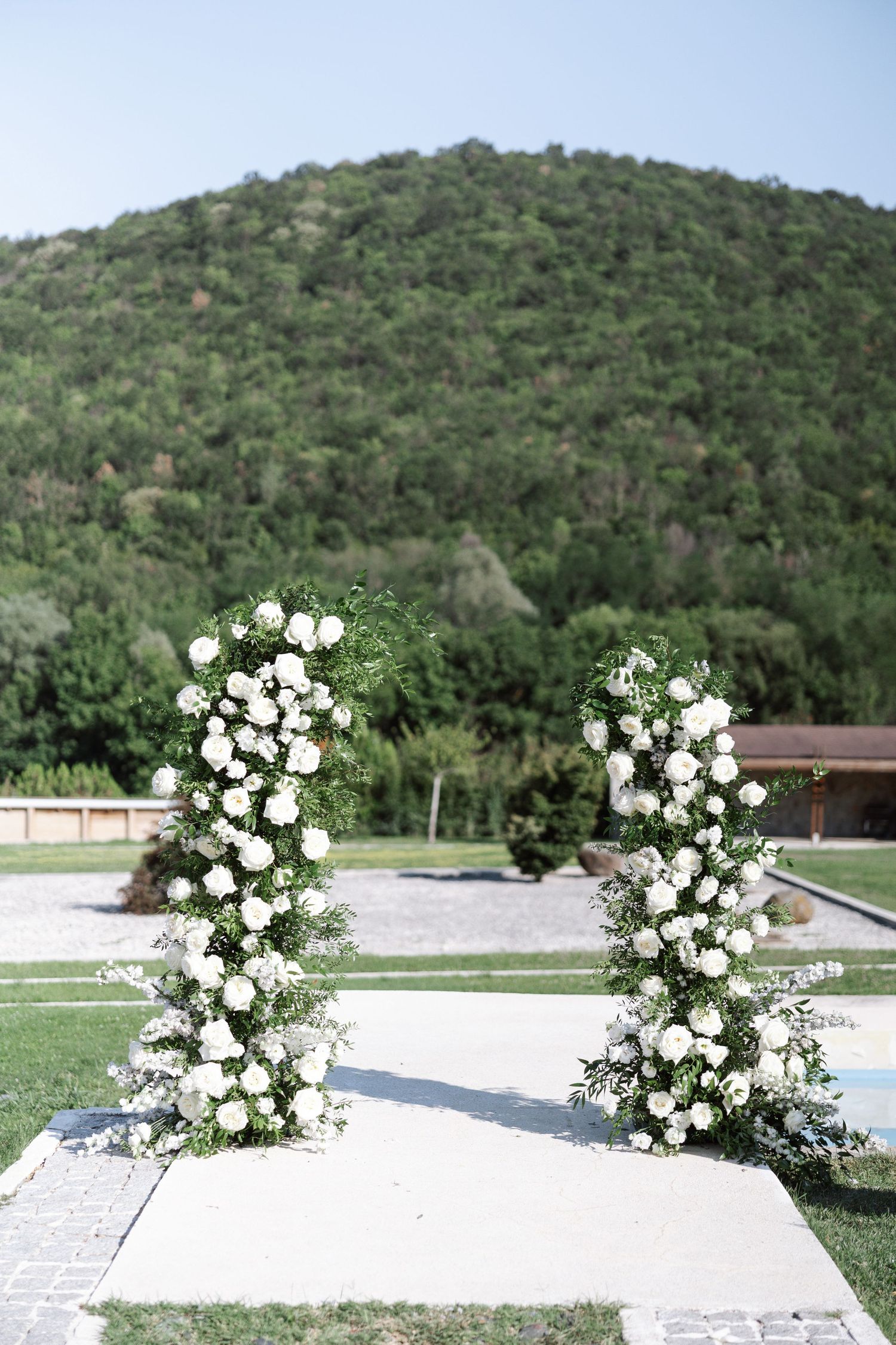 White floral arrangements adorn a row of marble headstones in a cemetery with green hills in the background.