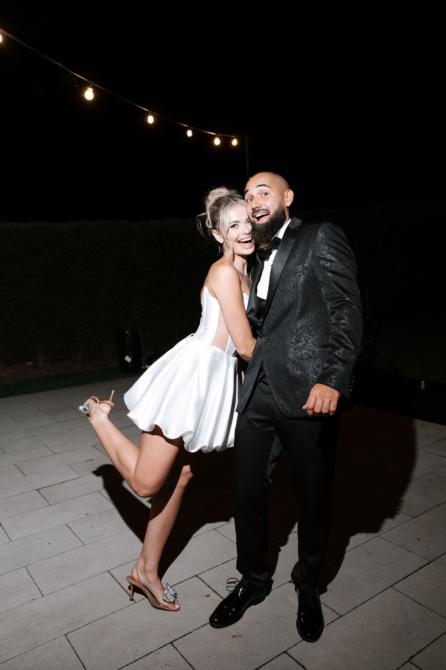 A couple shares a romantic dance dip moment at a nighttime outdoor wedding reception with string lights overhead.