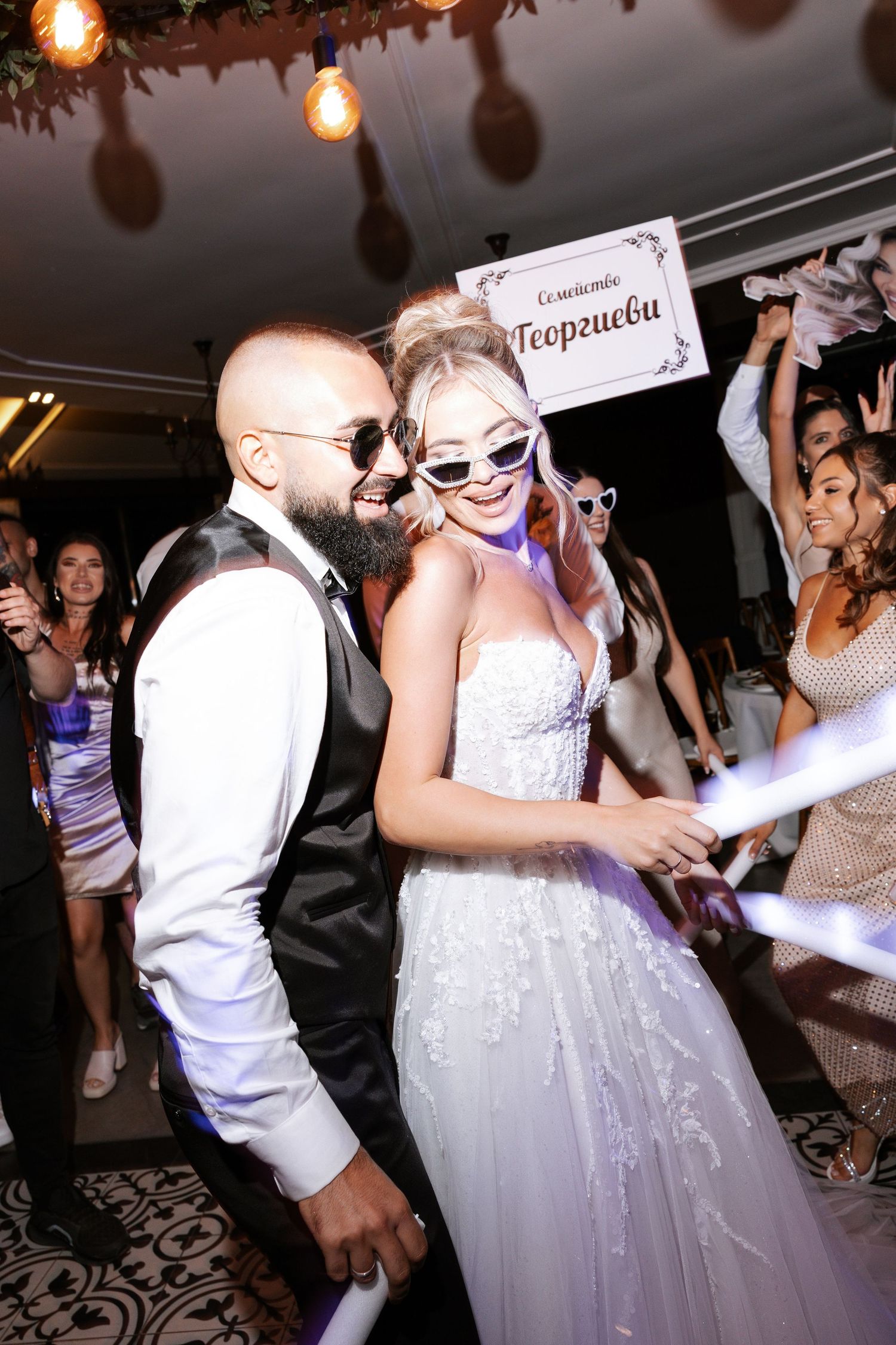 Wedding guests dance energetically on the dance floor at an evening reception with glowing lights overhead.