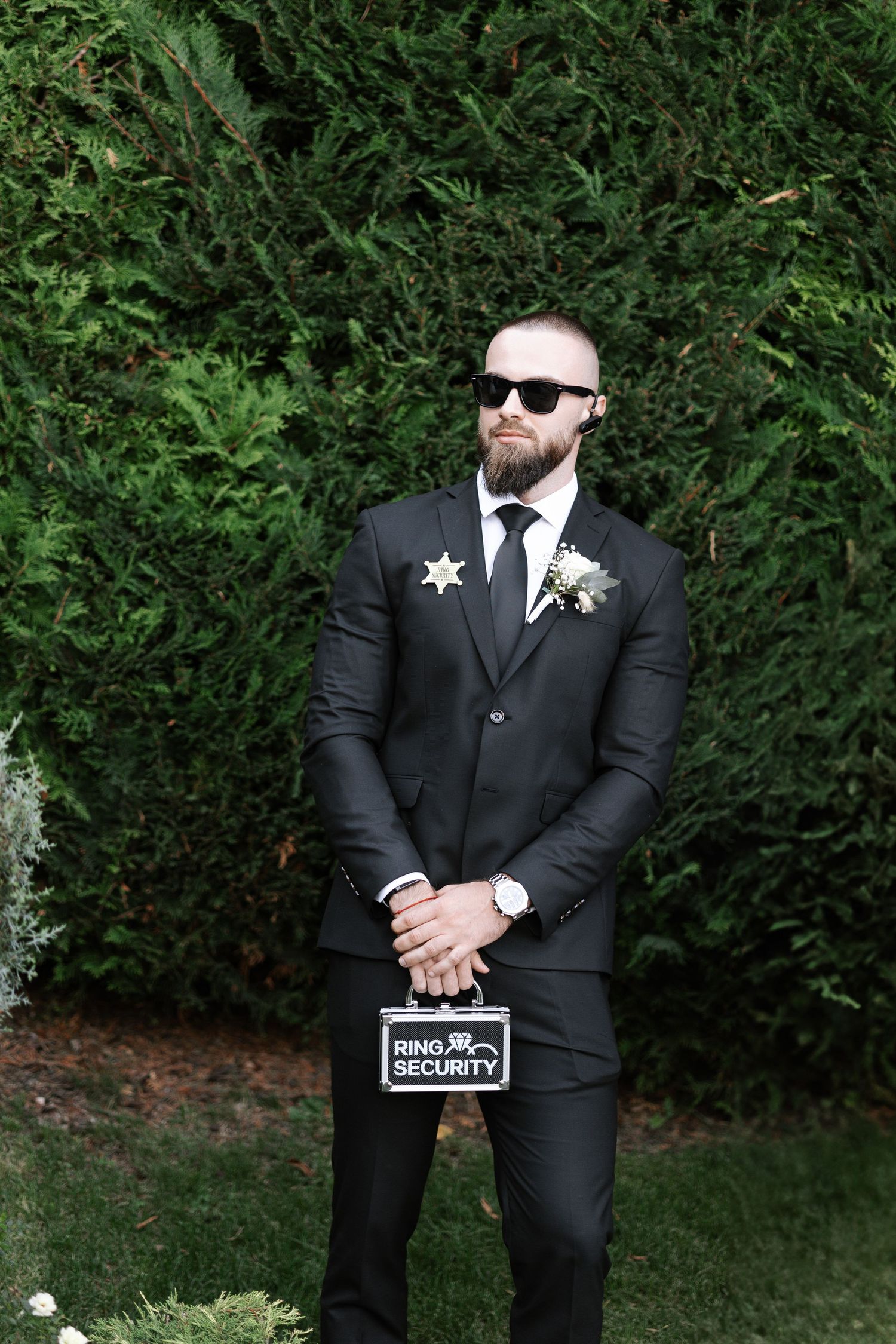 A person in a black suit and sunglasses poses in front of green hedges for wedding photos.