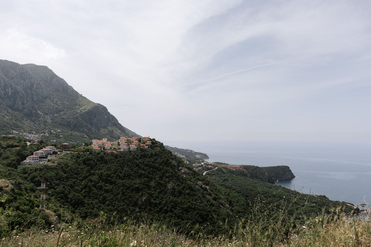Coastal village perched on green hillside overlooking Mediterranean Sea with rugged cliffs and cloudy sky.