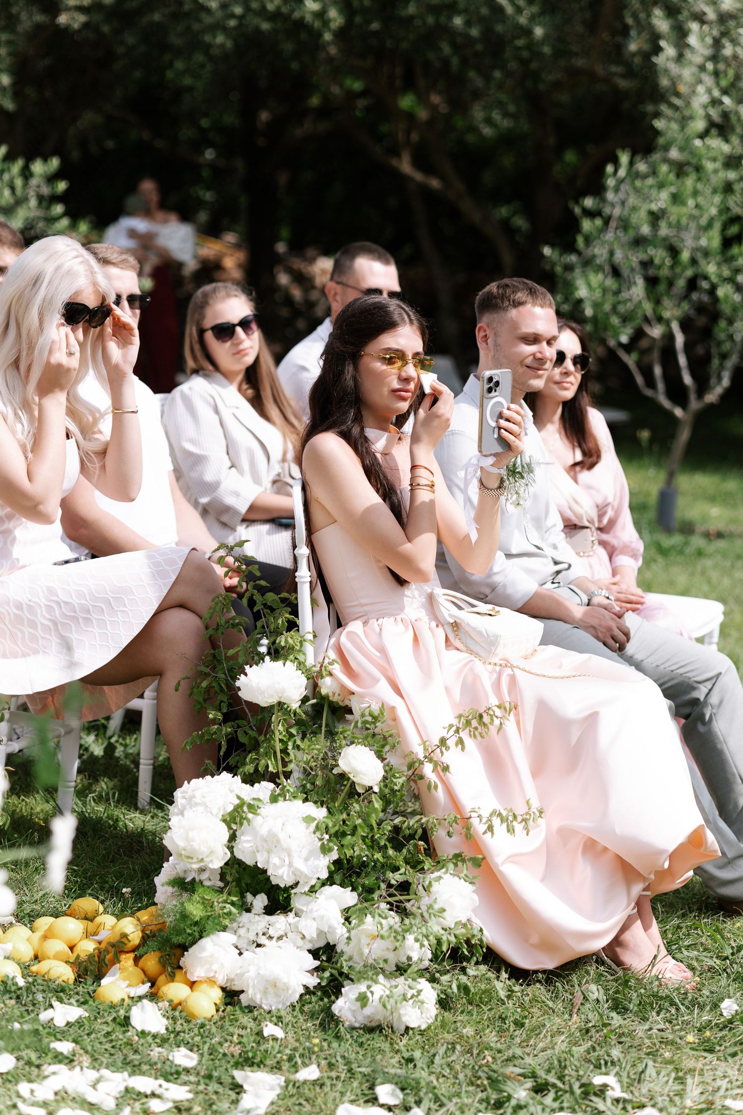 Wedding party guests in light pink dresses sit casually on grass with white hydrangea flowers at an outdoor garden event.