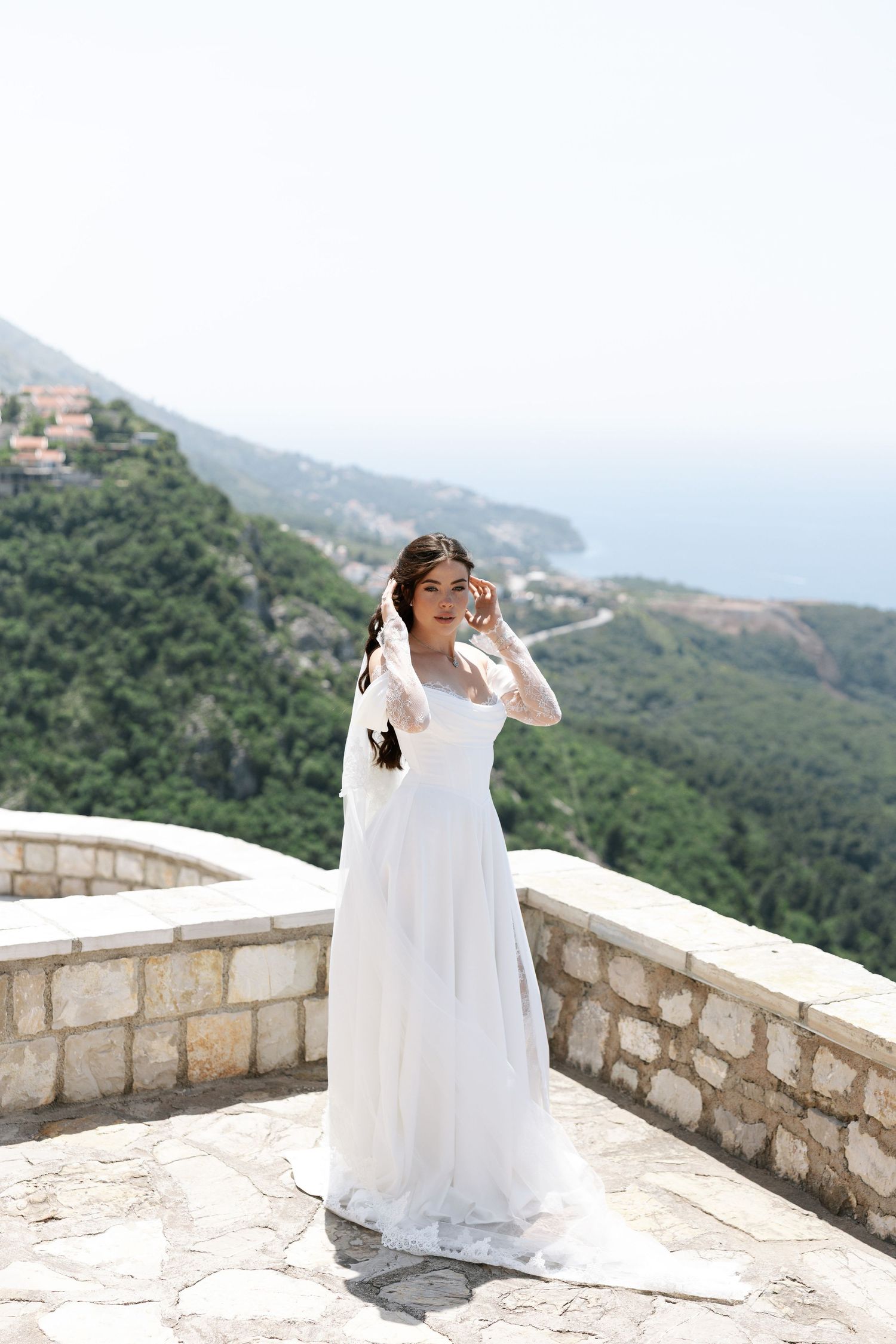 A woman in a flowing white dress poses gracefully on a stone balcony overlooking scenic mountain views.