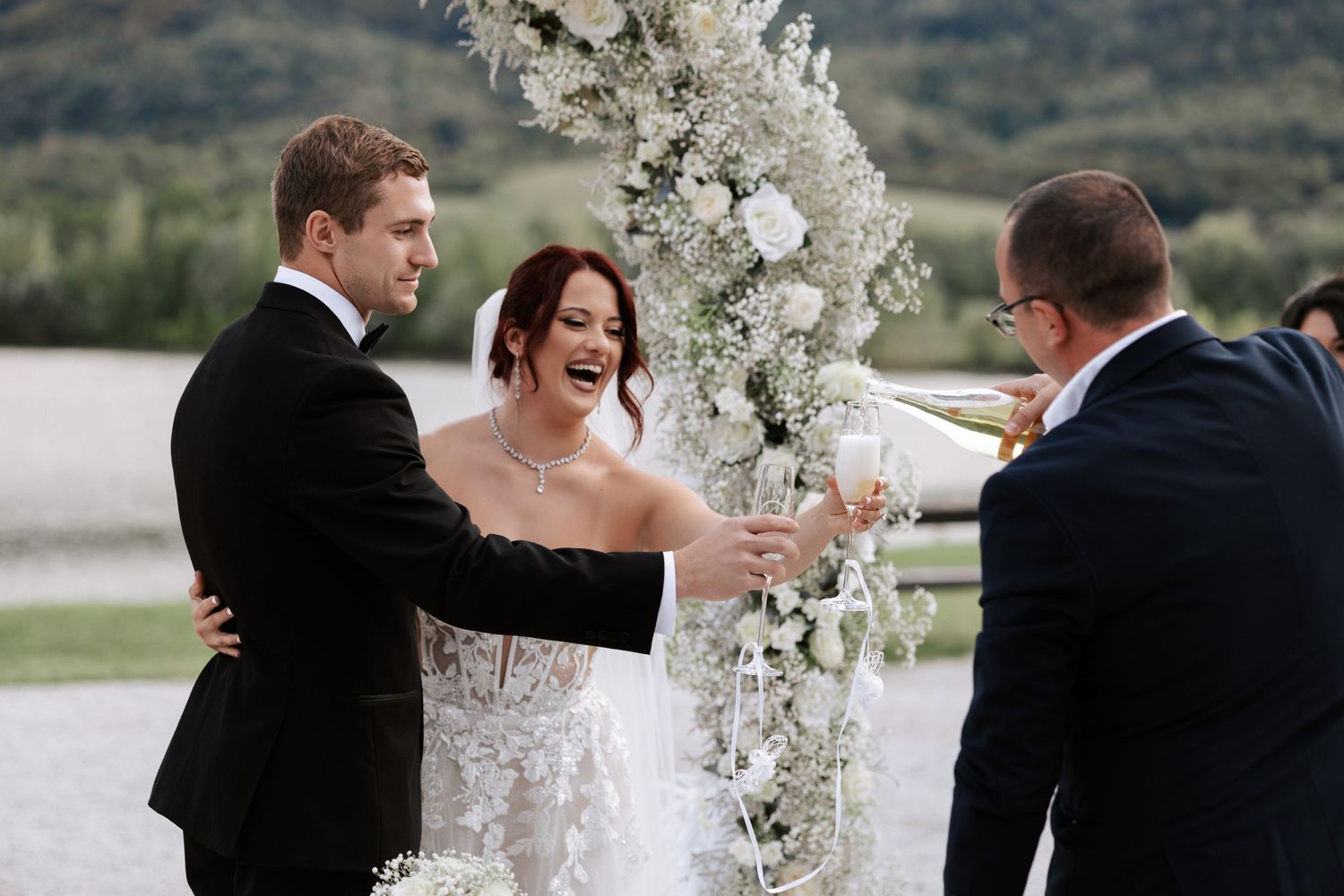 Wedding ceremony moment under white floral arch with officiant and mountain views.