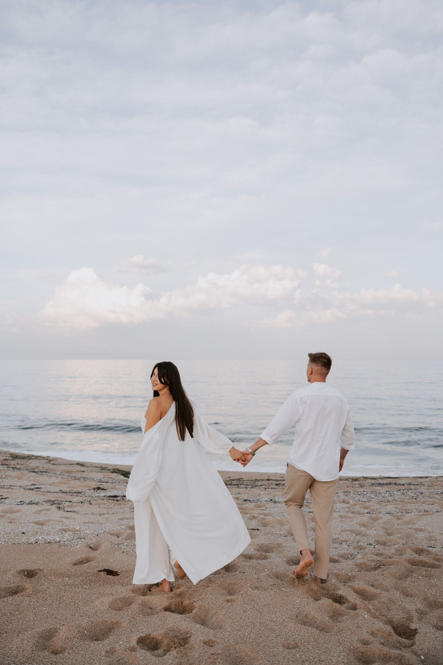 Couple in white clothing holds hands while walking on beach at sunset.