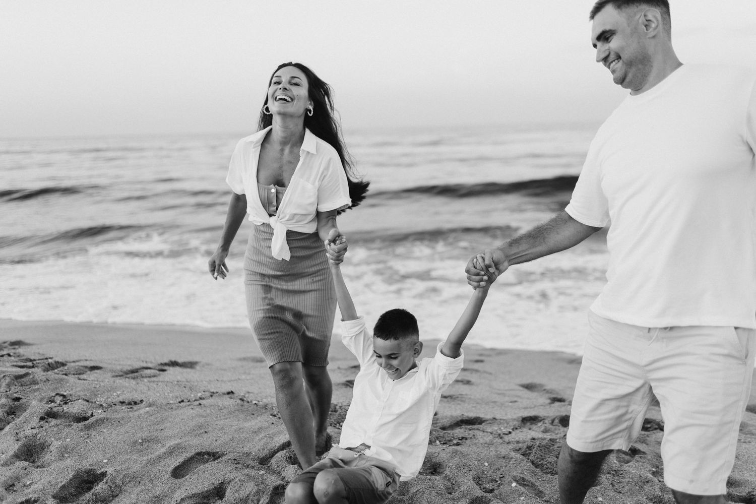 A happy family laughing and playing together on a sandy beach with ocean waves.