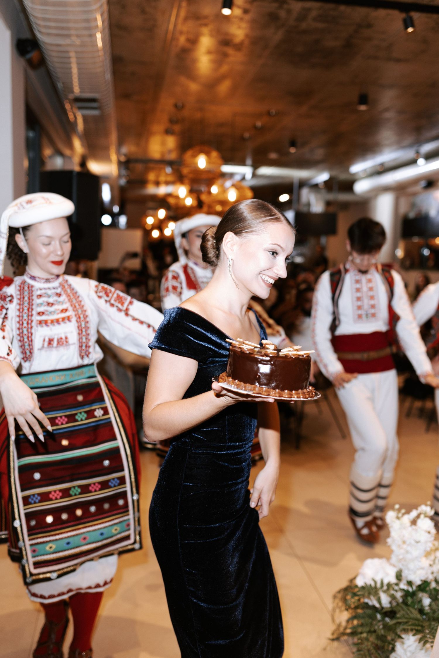 Performers in traditional folk costumes carrying a chocolate cake through an indoor venue.