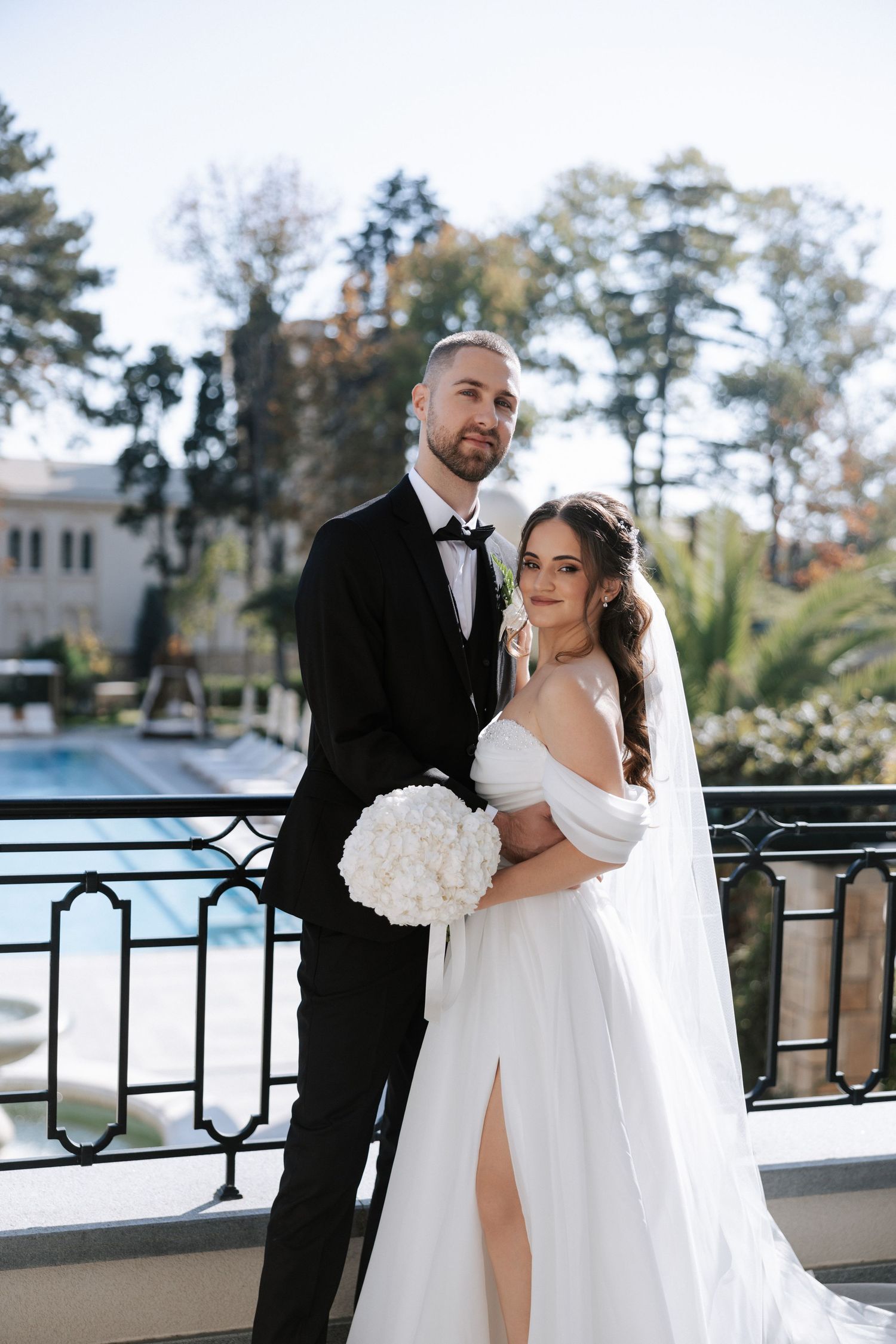 Elegant couple in wedding attire poses on balcony with swimming pool and architecture in background.
