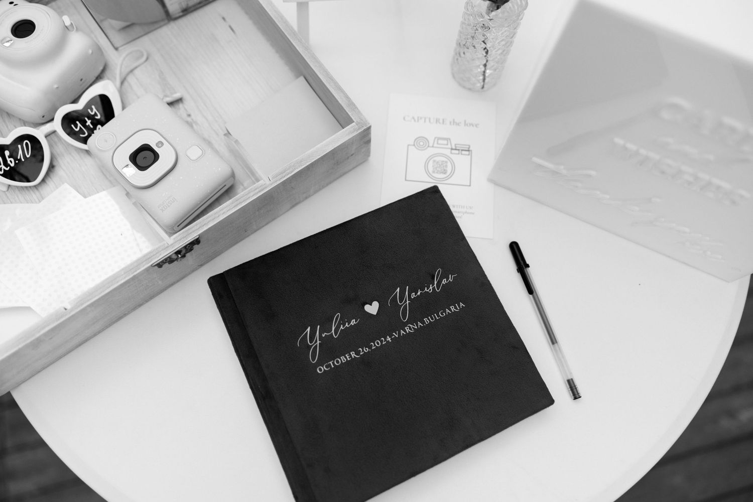 Black wedding guestbook and pen on white surface with scattered planning materials.