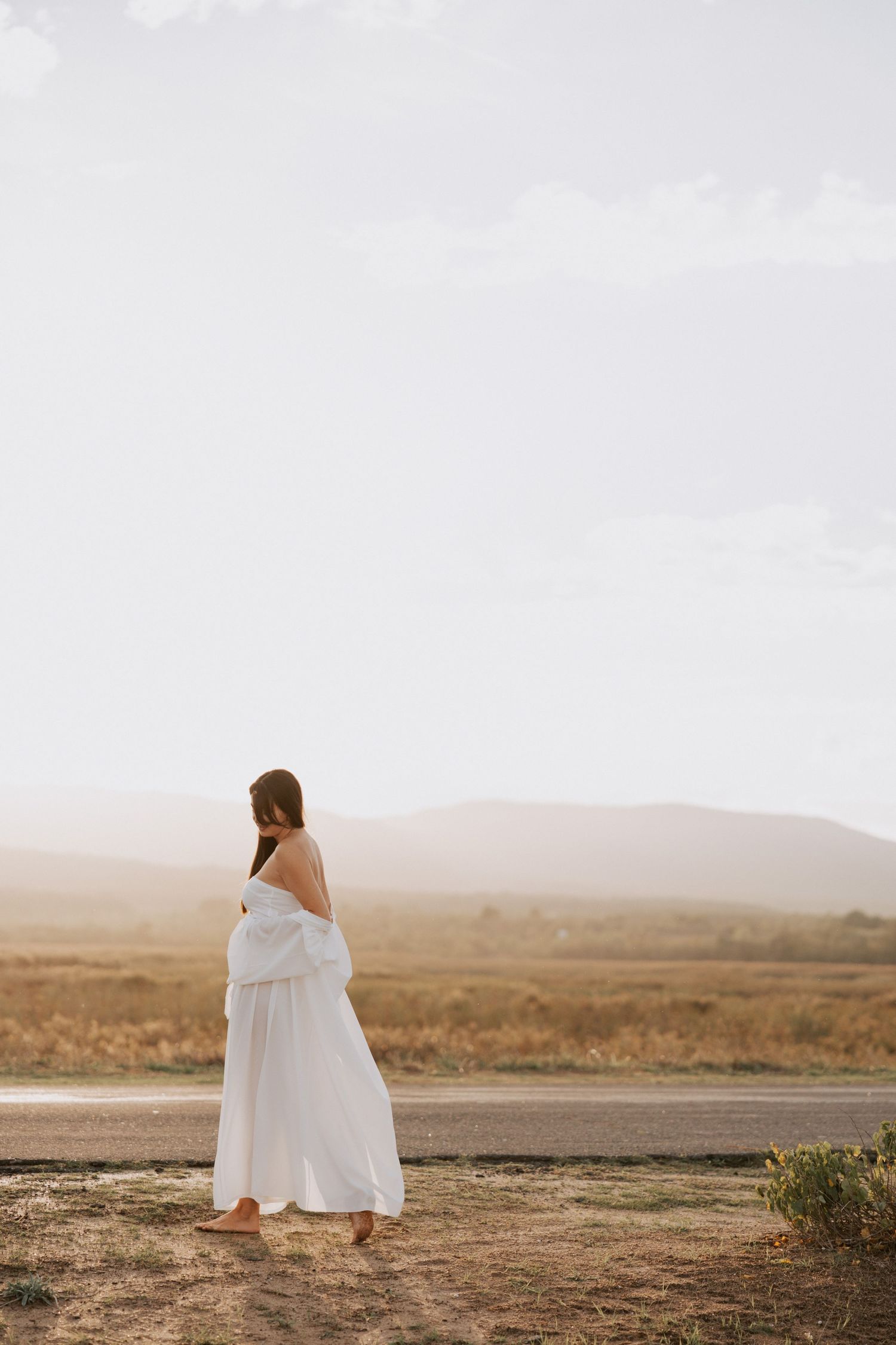 Woman in ethereal white dress stands barefoot overlooking desert landscape at sunset.