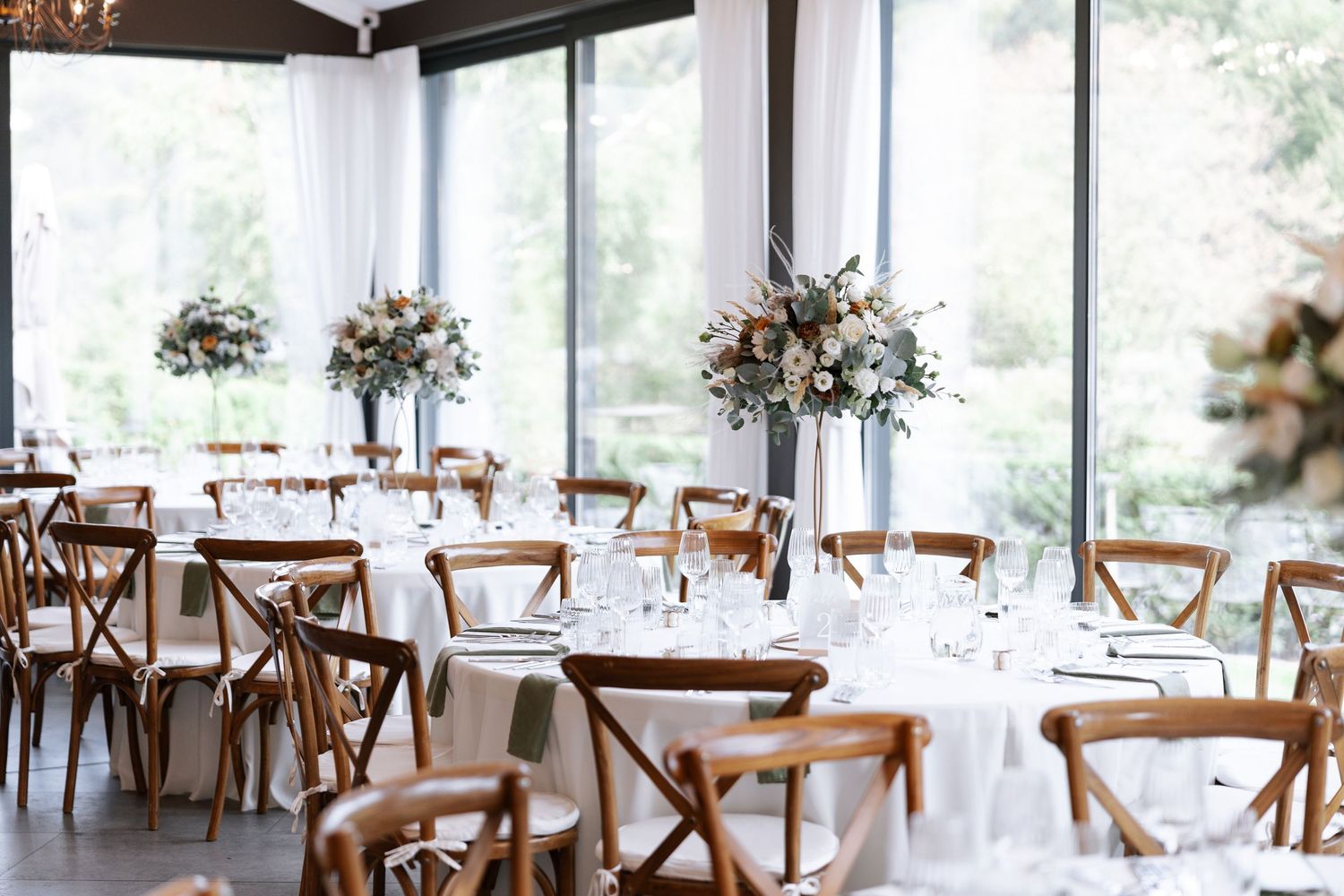 Elegant wedding reception venue with wooden cross-back chairs and round tables set in a bright, window-lined space.
