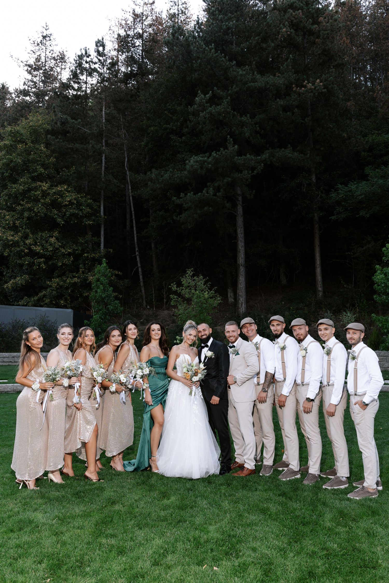 Wedding party poses in line formation on green lawn with bridesmaids in beige and groomsmen in light colored suits.