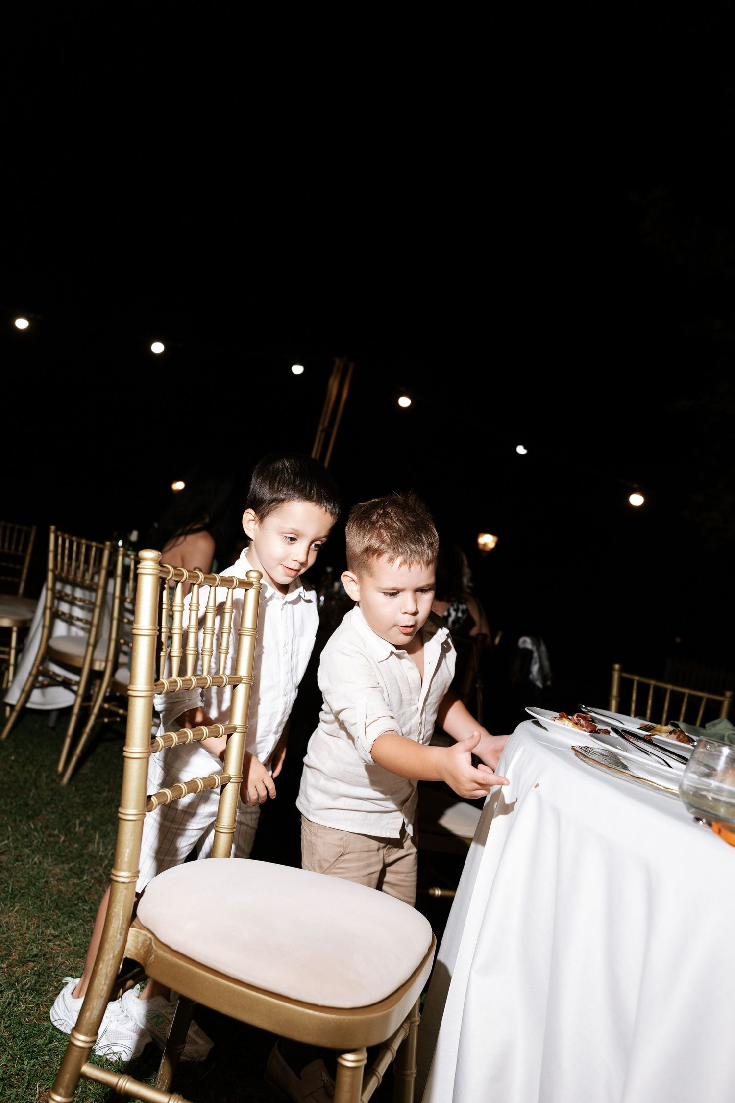Young children in formal attire sit at a white-clothed table during an evening outdoor reception.
