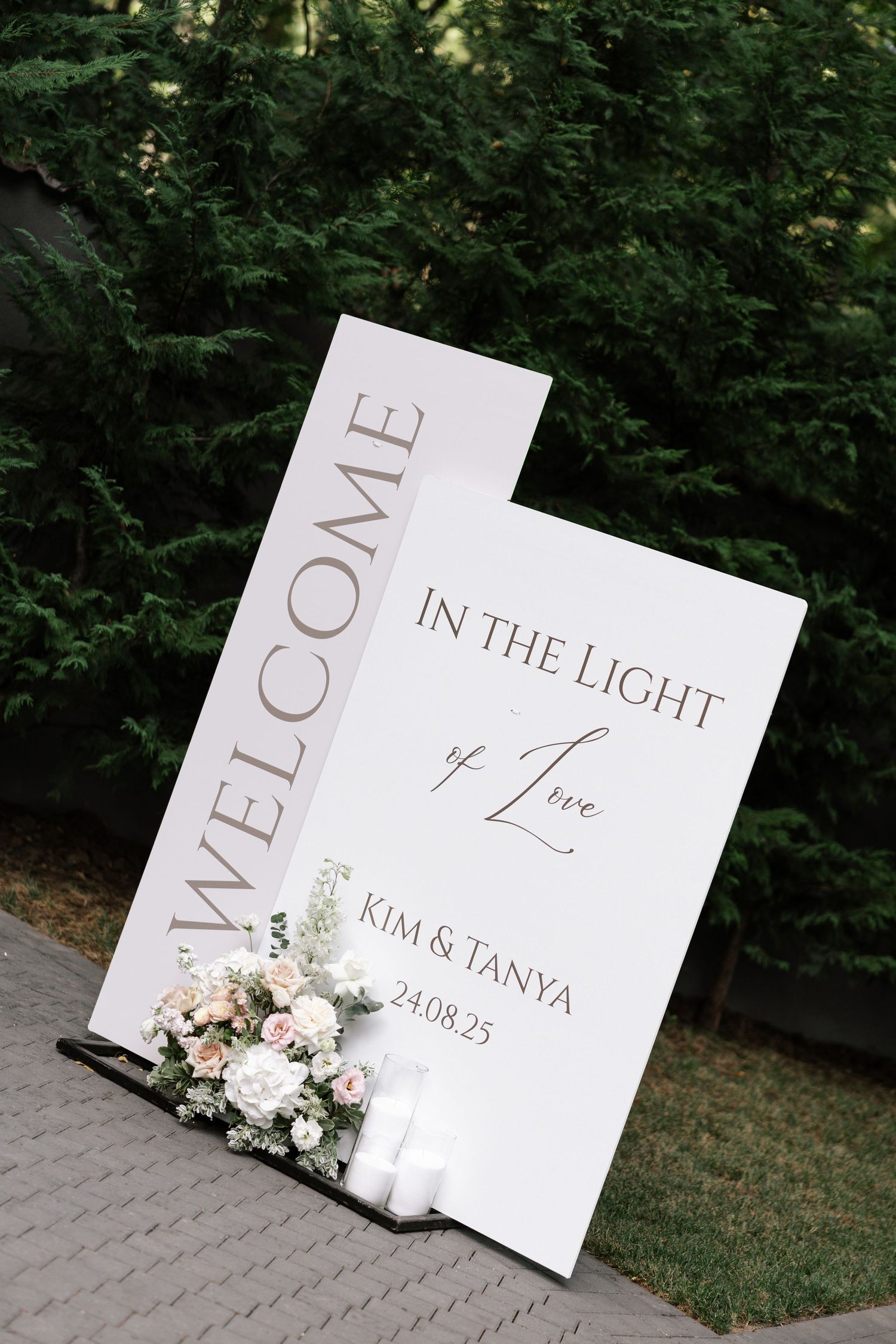 Modern white wedding welcome sign with floral decorations against a green outdoor backdrop.