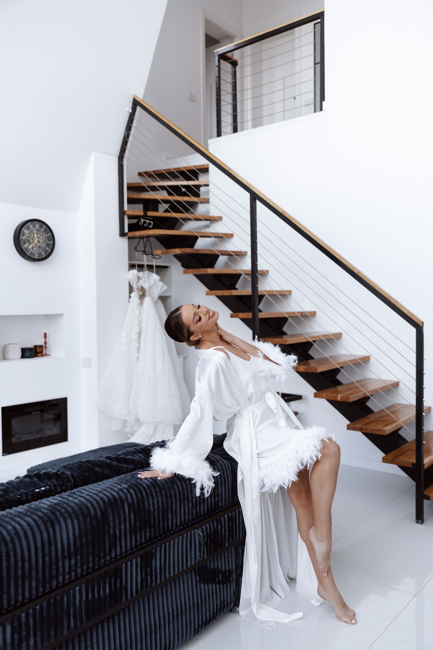 A figure in a flowing white dress poses gracefully on modern wooden stairs with black railing in a minimalist interior.