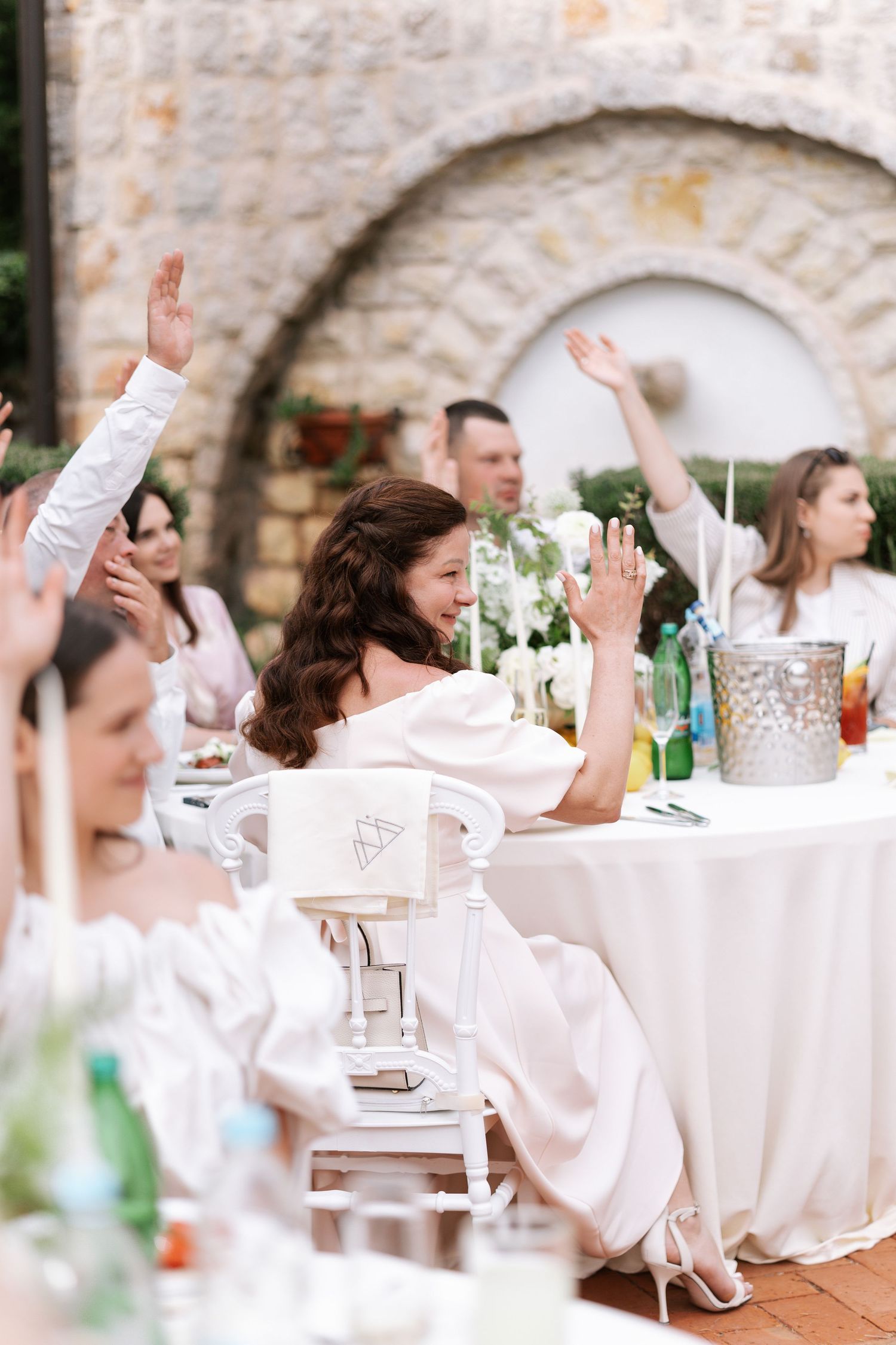 Wedding guests raise their hands in celebration at an outdoor reception with white tablecloths and stone walls.
