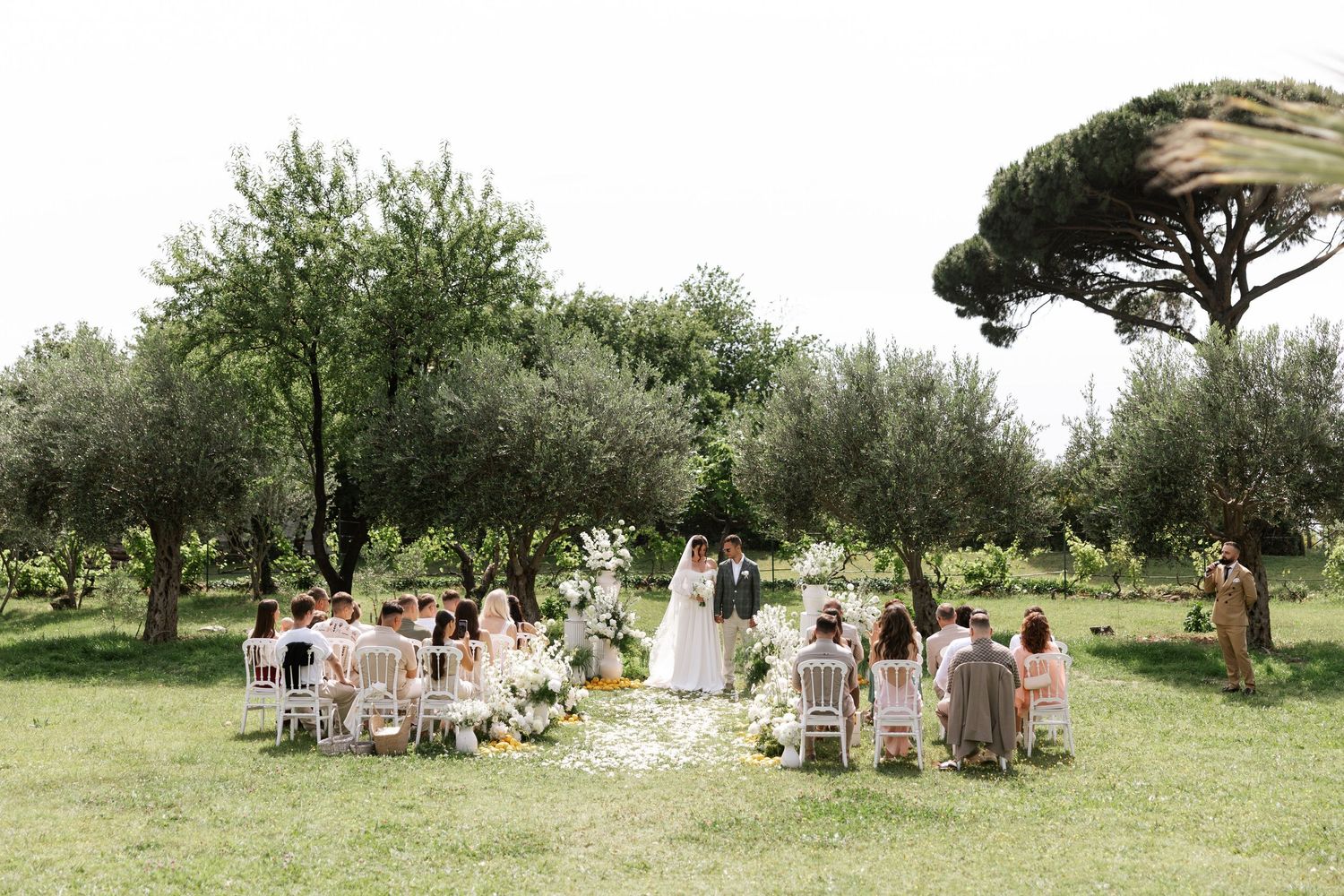 Outdoor wedding ceremony setup with white chairs arranged in rows beneath olive trees in a rustic garden setting.