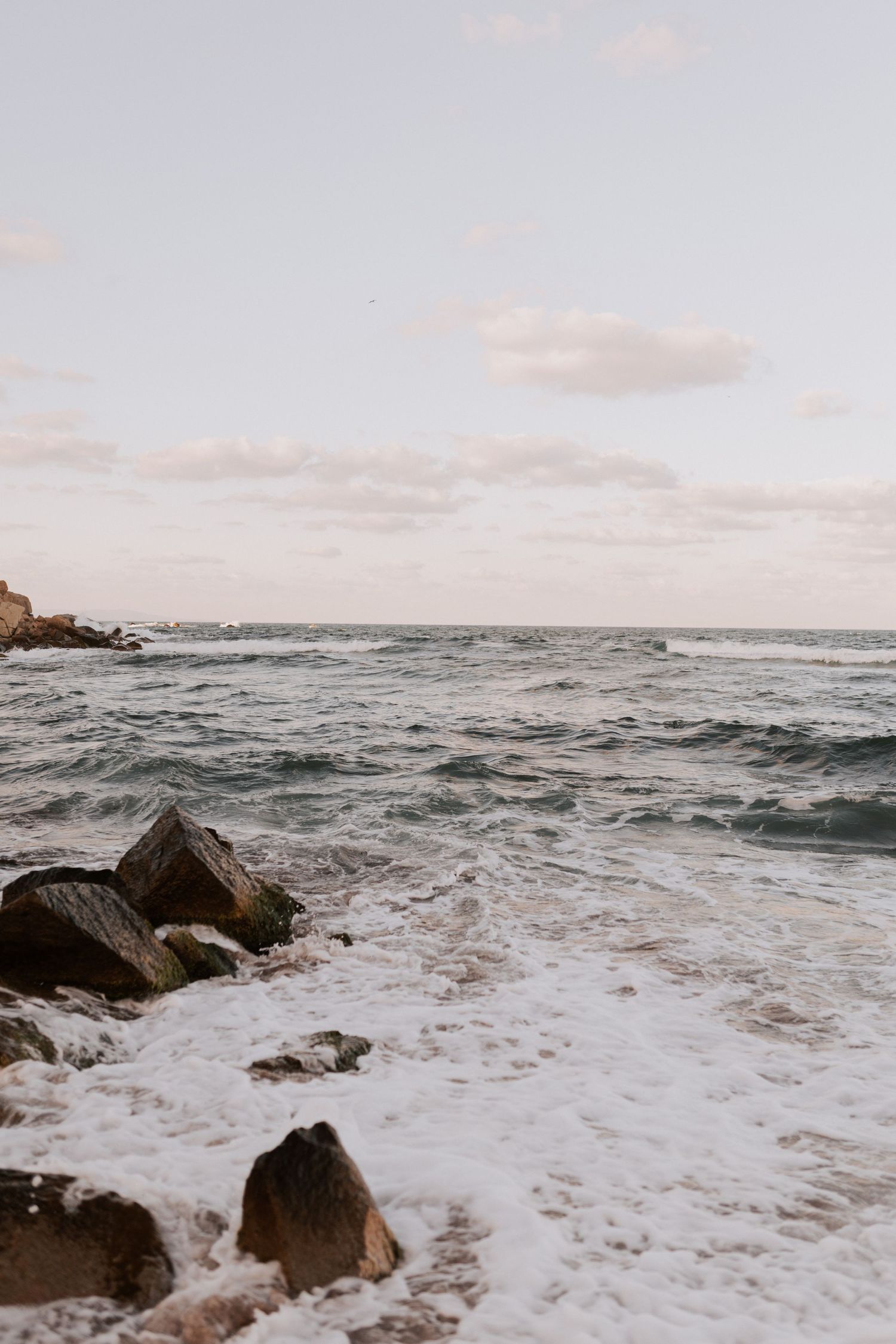 Dramatic ocean waves crash against dark rocks along a rugged coastline at sunset.