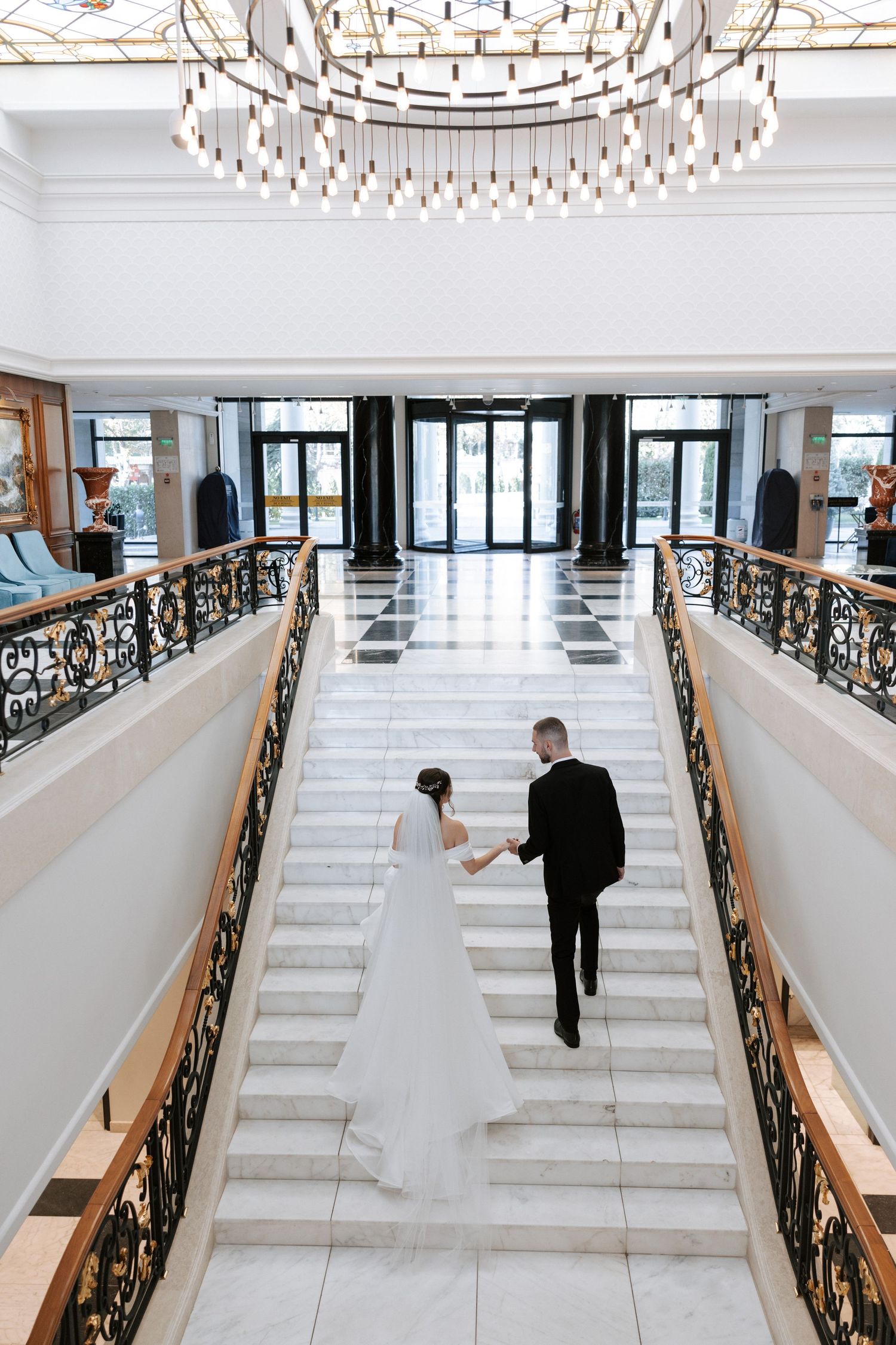 Aerial view of newlyweds walking down grand marble staircase under crystal chandelier.