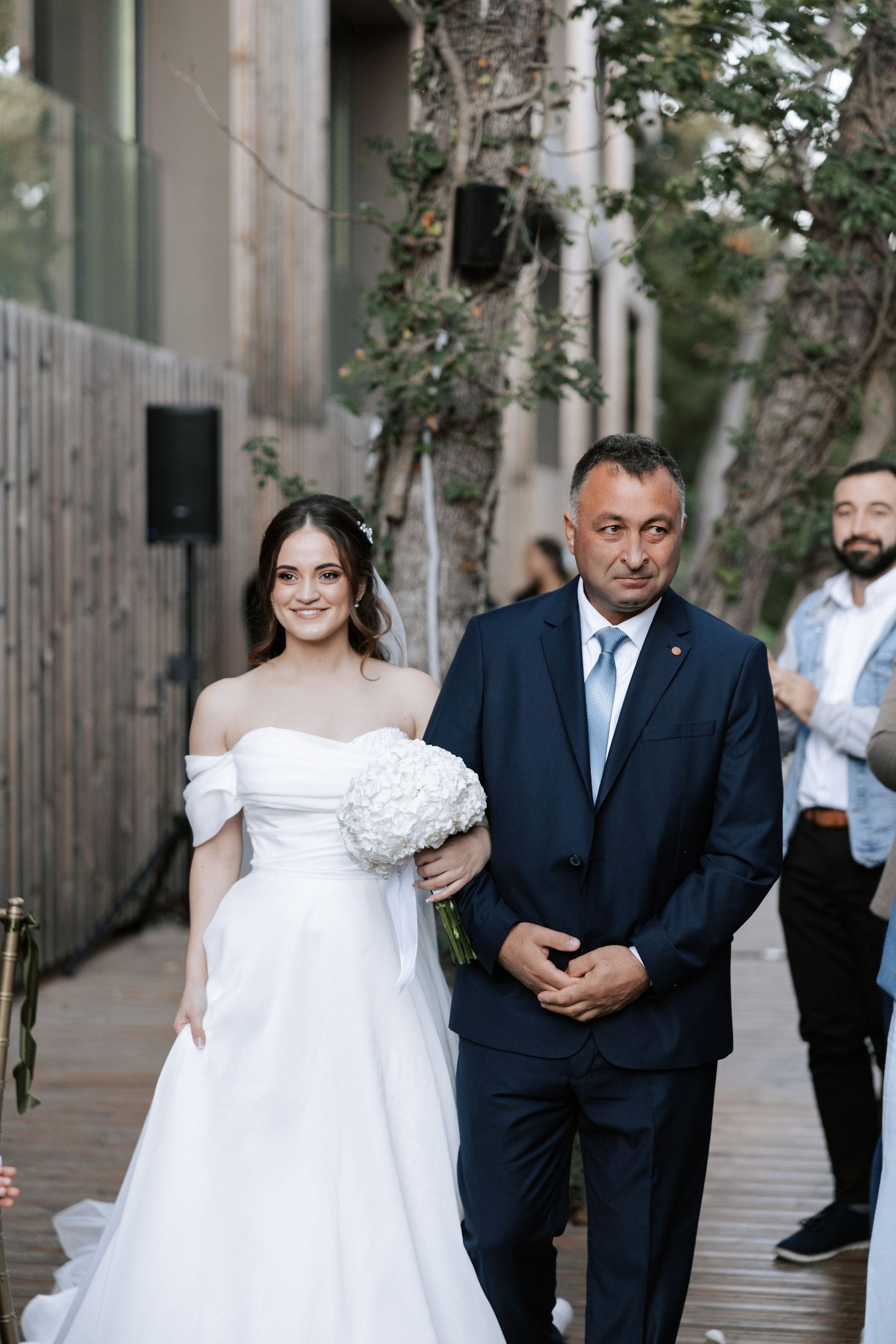 A wedding ceremony procession takes place on a wooden deck with trees and modern architecture.