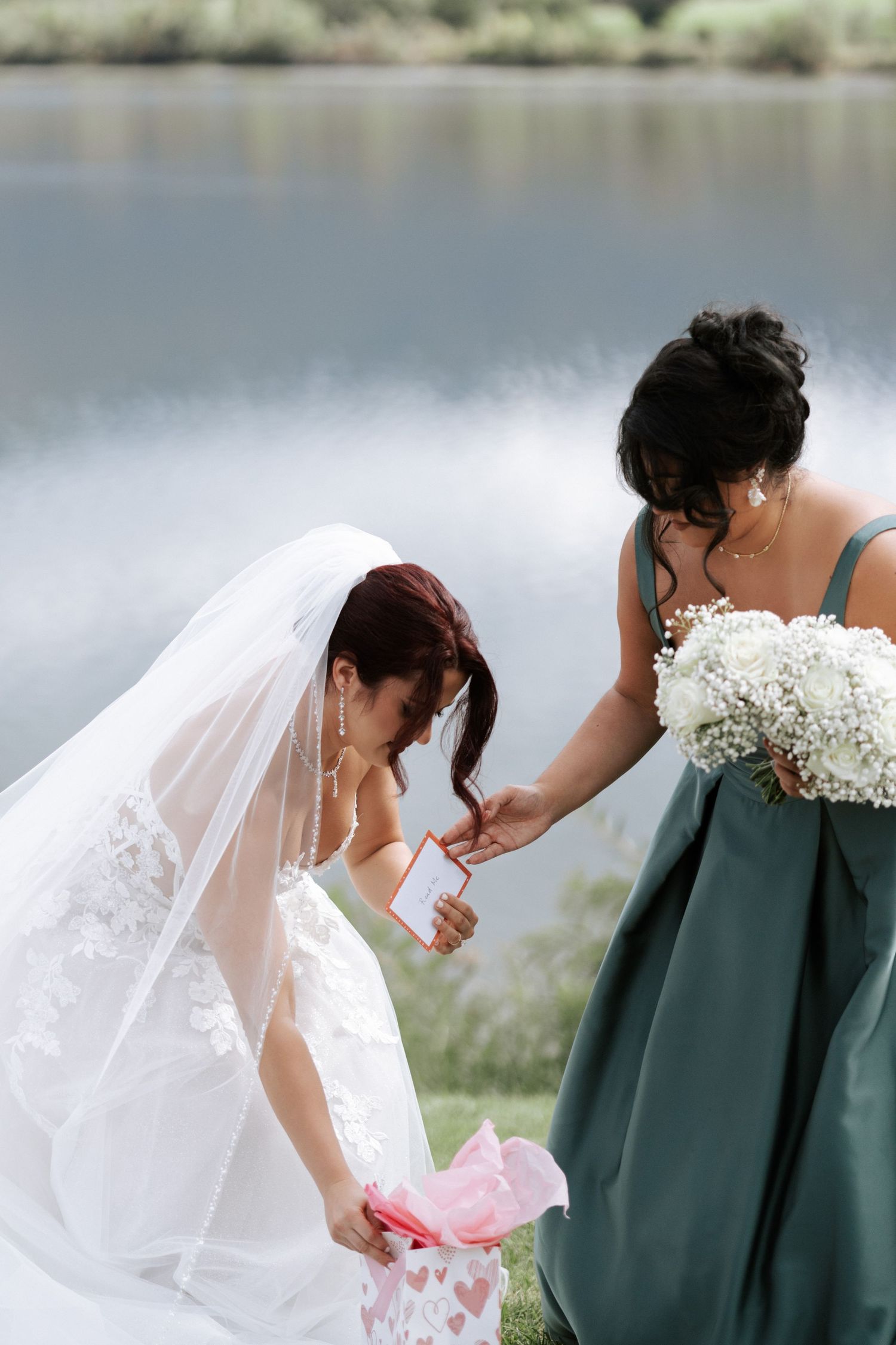 A bride in white lace gown opens gifts beside attendant in sage green dress by a serene lake.