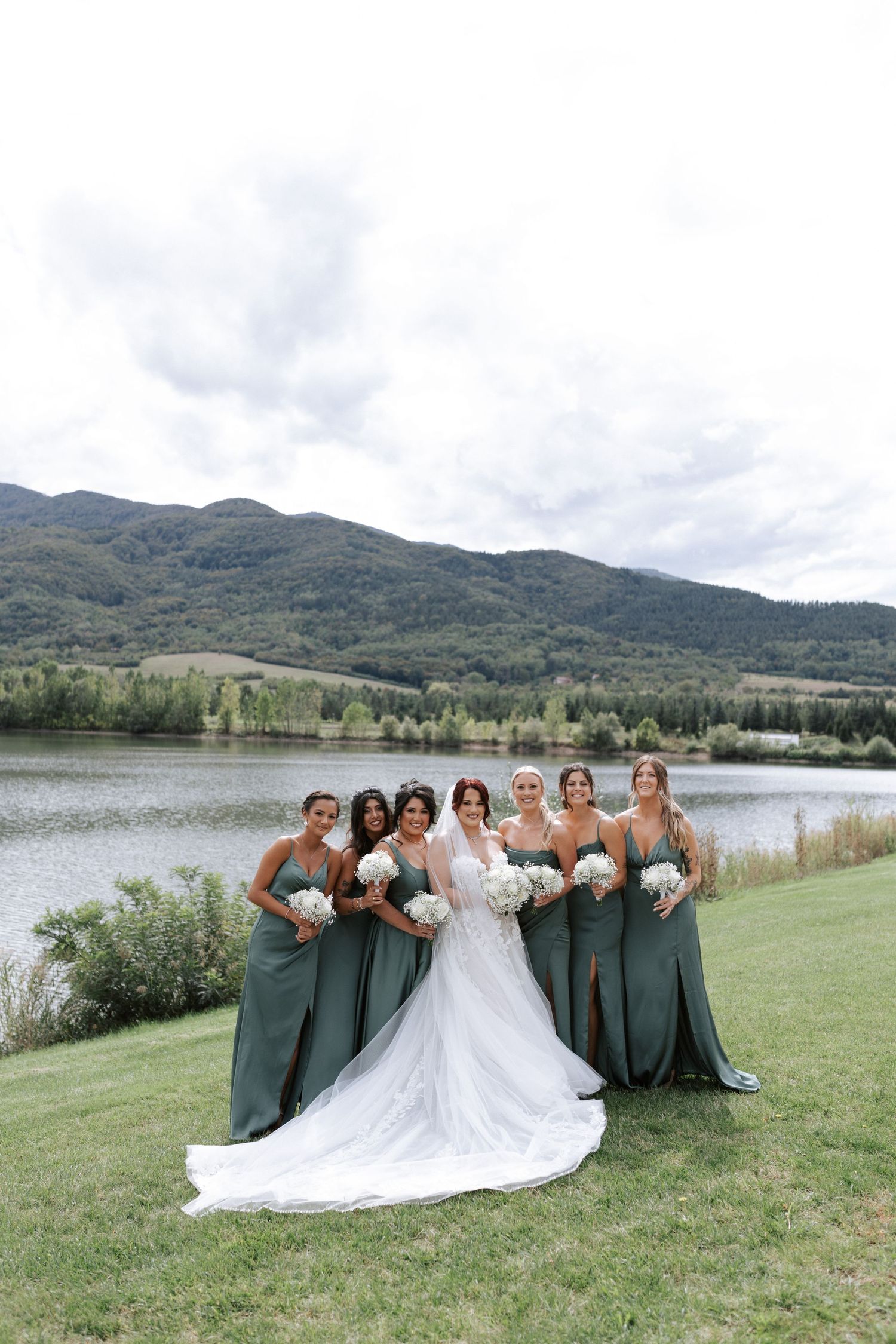 Wedding party in sage green dresses poses for formal photo against mountain lake backdrop.
