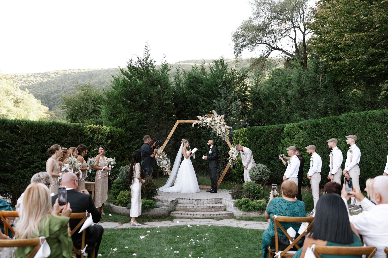 Wedding guests gather outdoors in a garden setting for a celebration surrounded by lush greenery and scenic views.