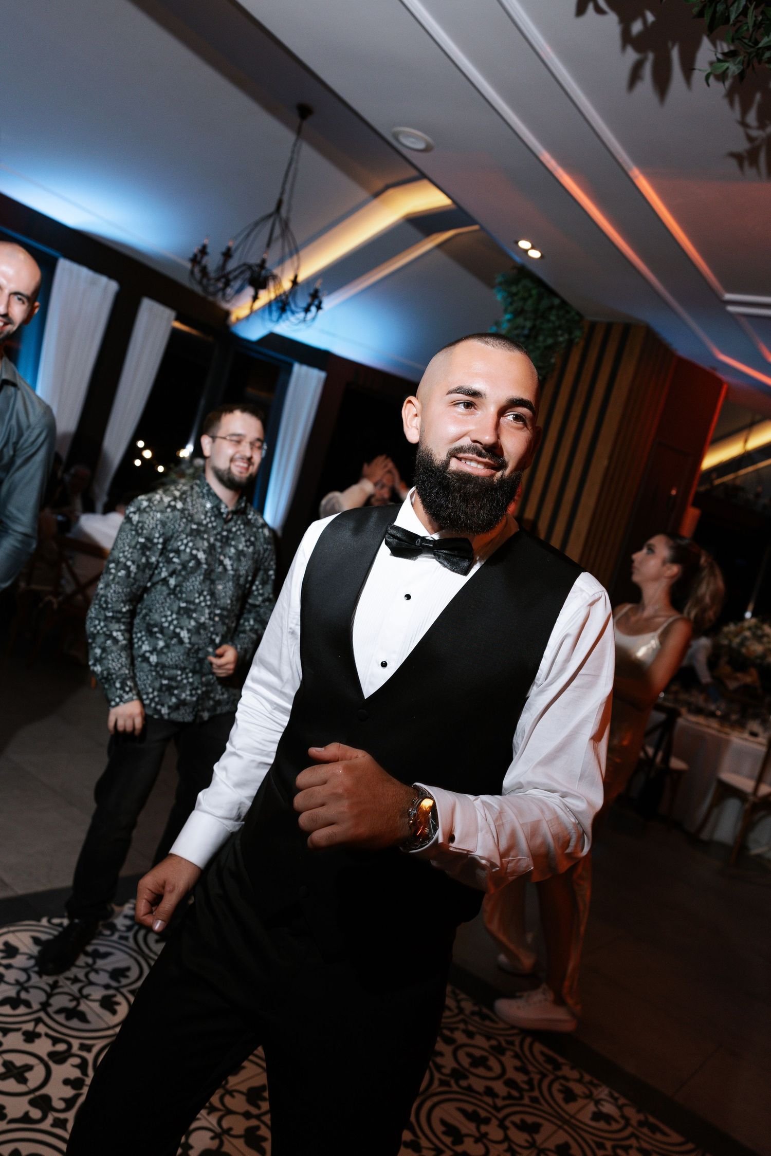 A well-dressed guest in formal attire with a black vest and bowtie smiles at a wedding reception.