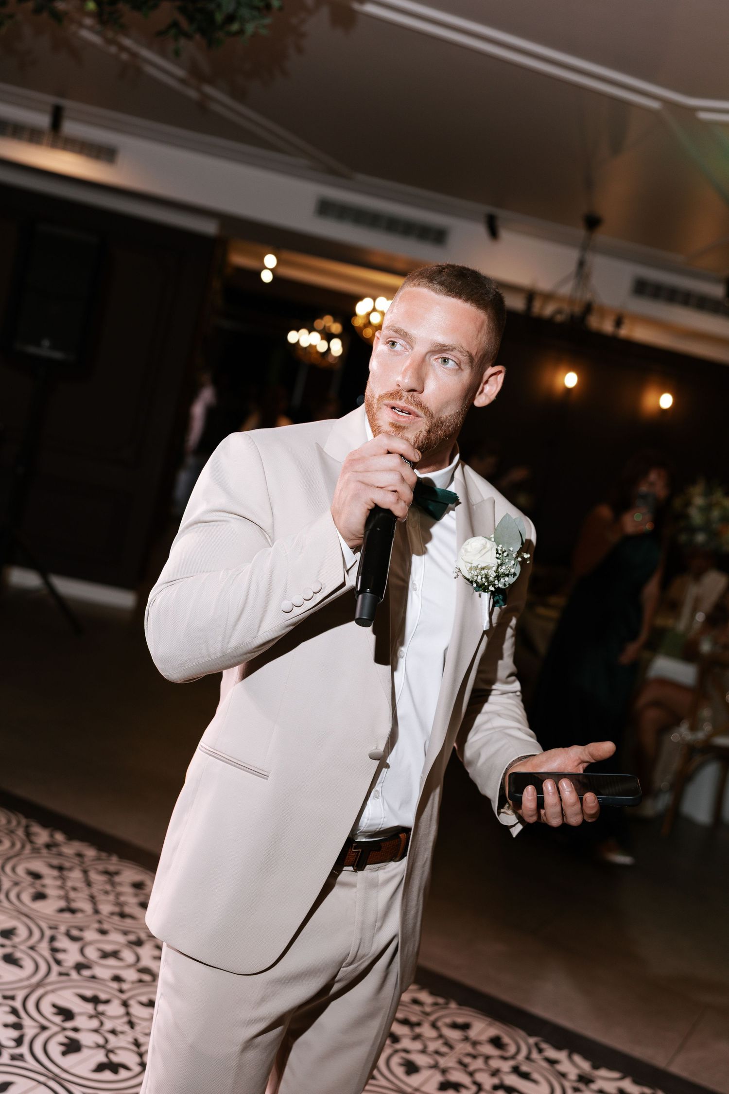 A person in a white suit and boutonniere gestures while dancing at an indoor reception venue.