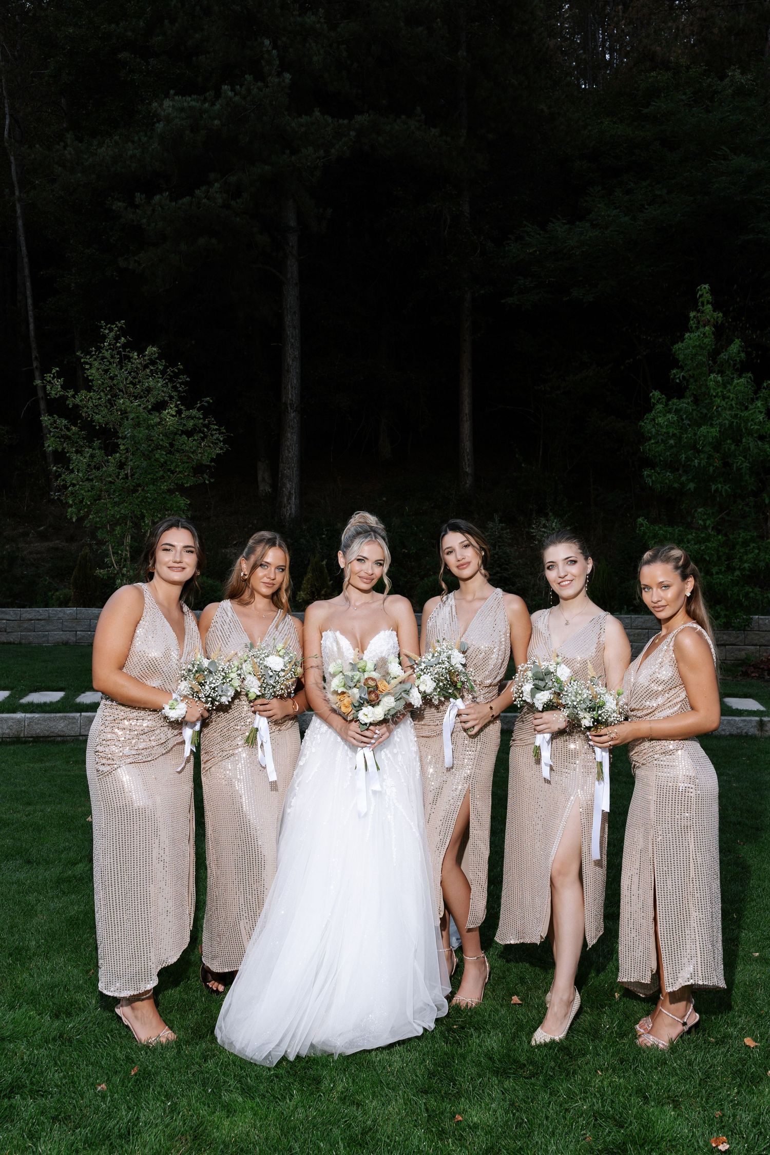 A bridal party in blush pink dresses and a bride in white pose together on a grassy lawn at dusk.