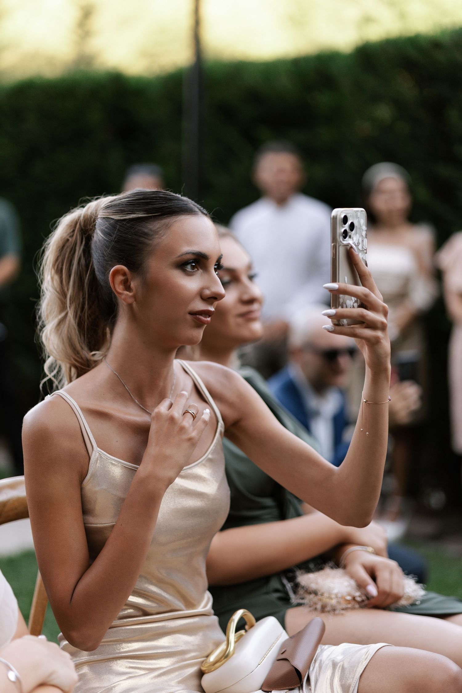 A group of people in formal attire sit outdoors while taking photos with their phones at an event.