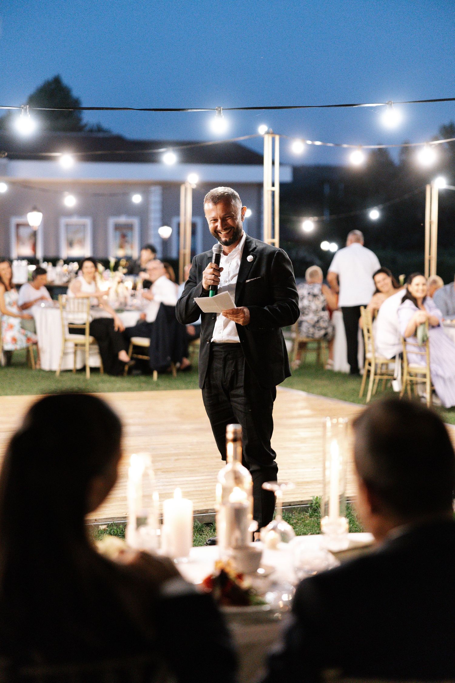 Outdoor evening wedding reception with string lights illuminating guests mingling under the night sky.