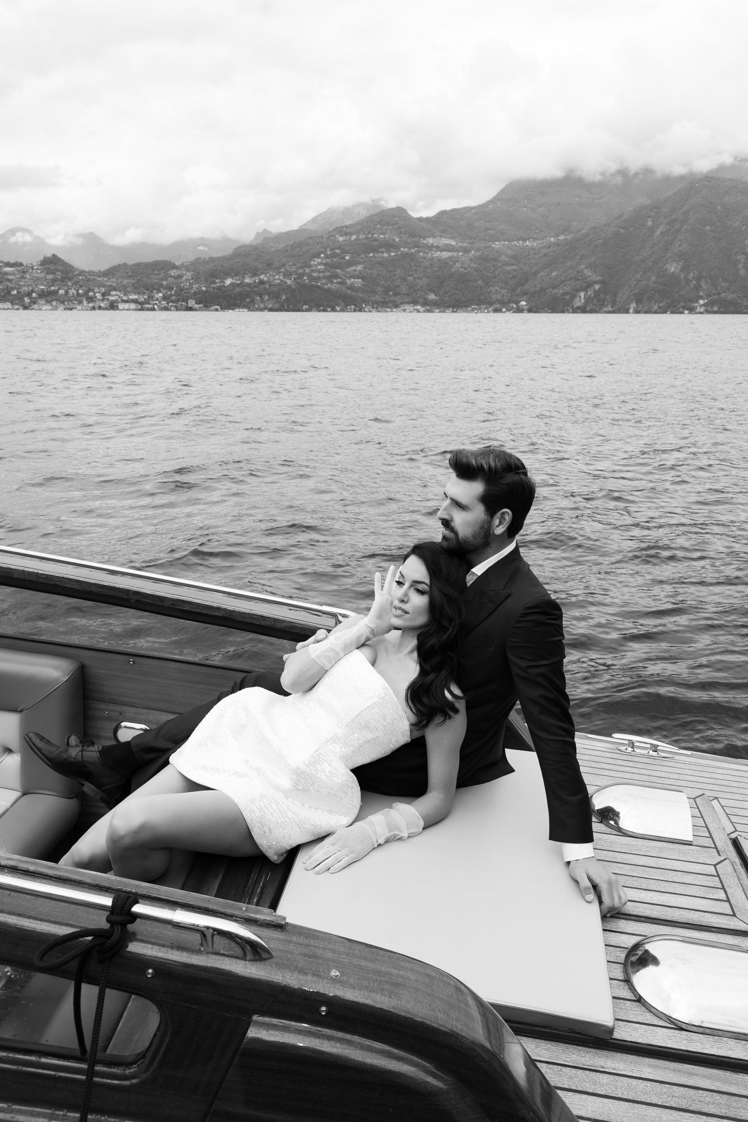 A couple relaxes on a wooden boat deck with mountains and lake scenery in the background.