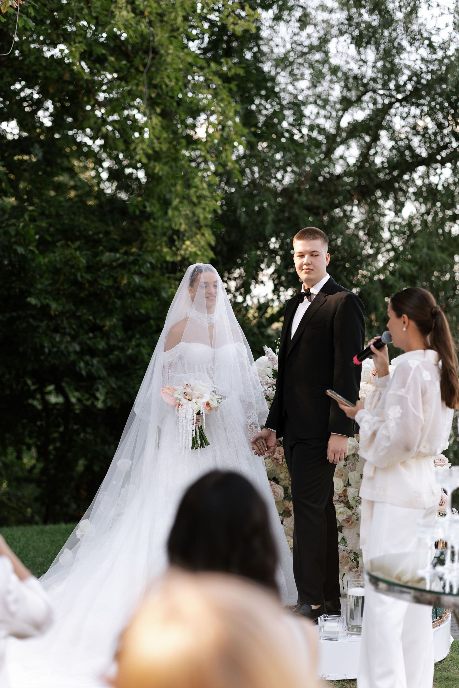 An outdoor wedding ceremony with white decor and floral arrangements against a backdrop of trees.