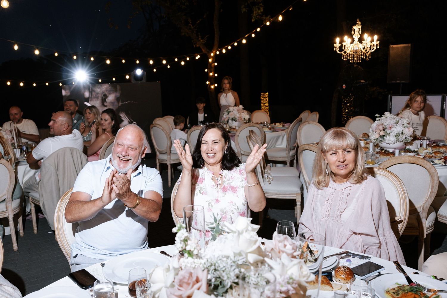 Wedding reception guests enjoying dinner under string lights and chandelier.