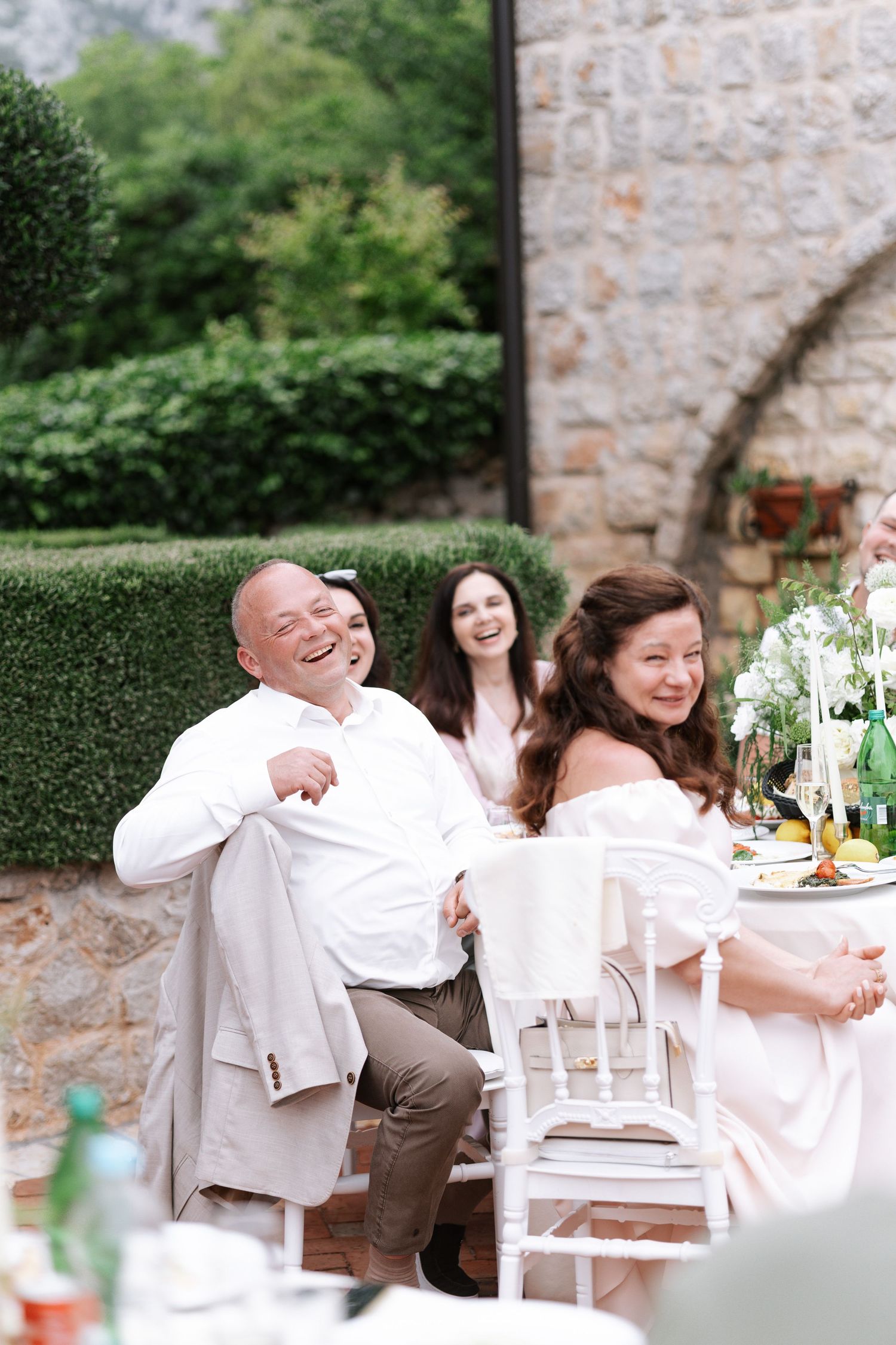 Wedding guests share joyful laughter at an elegant outdoor reception with stone walls and greenery in the background.