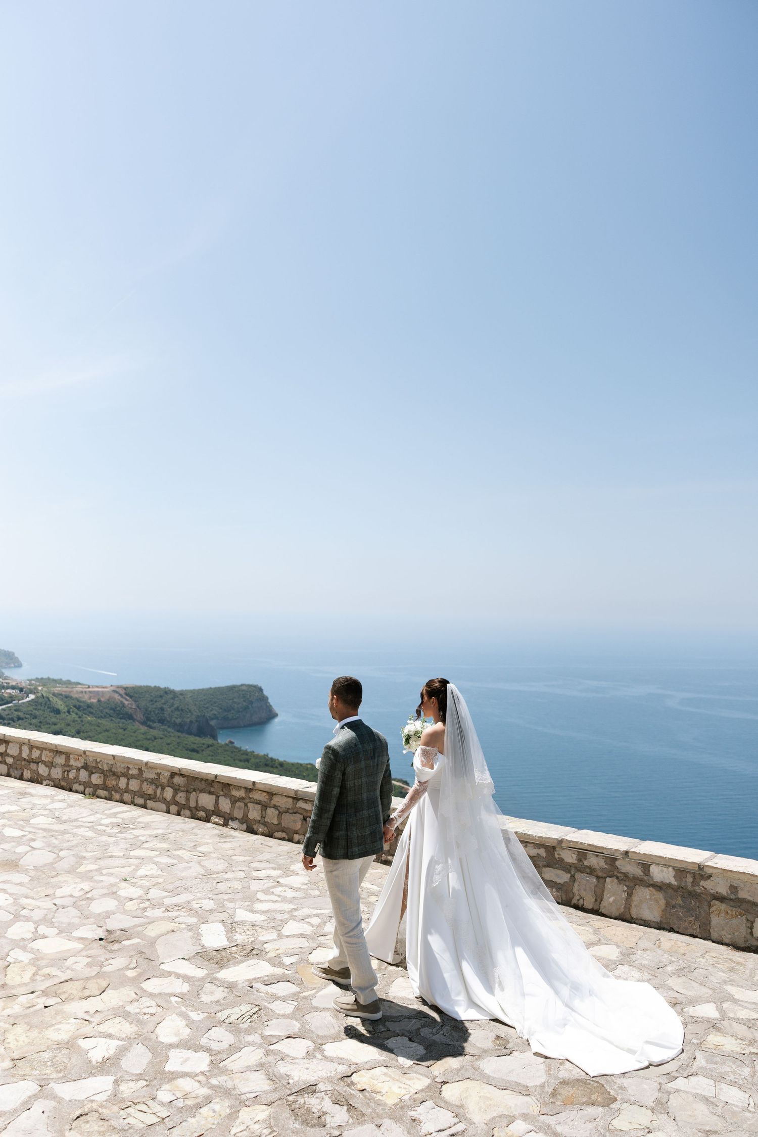 A couple in wedding attire stands on a stone terrace overlooking a scenic coastal view on their special day.