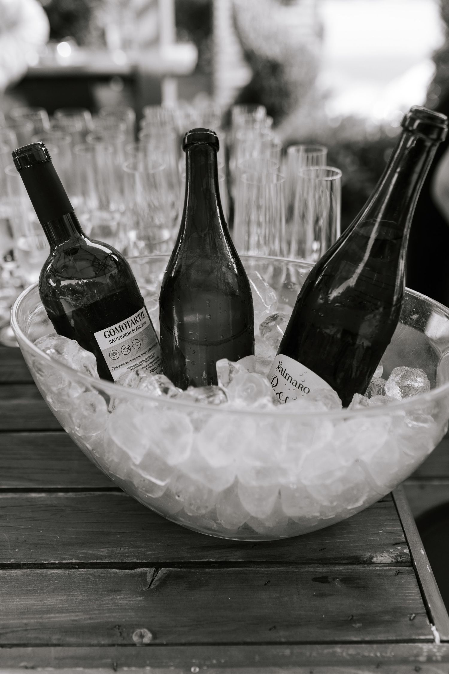 Black and white photo of wine bottles chilling in a bowl of ice with empty glasses in the background.