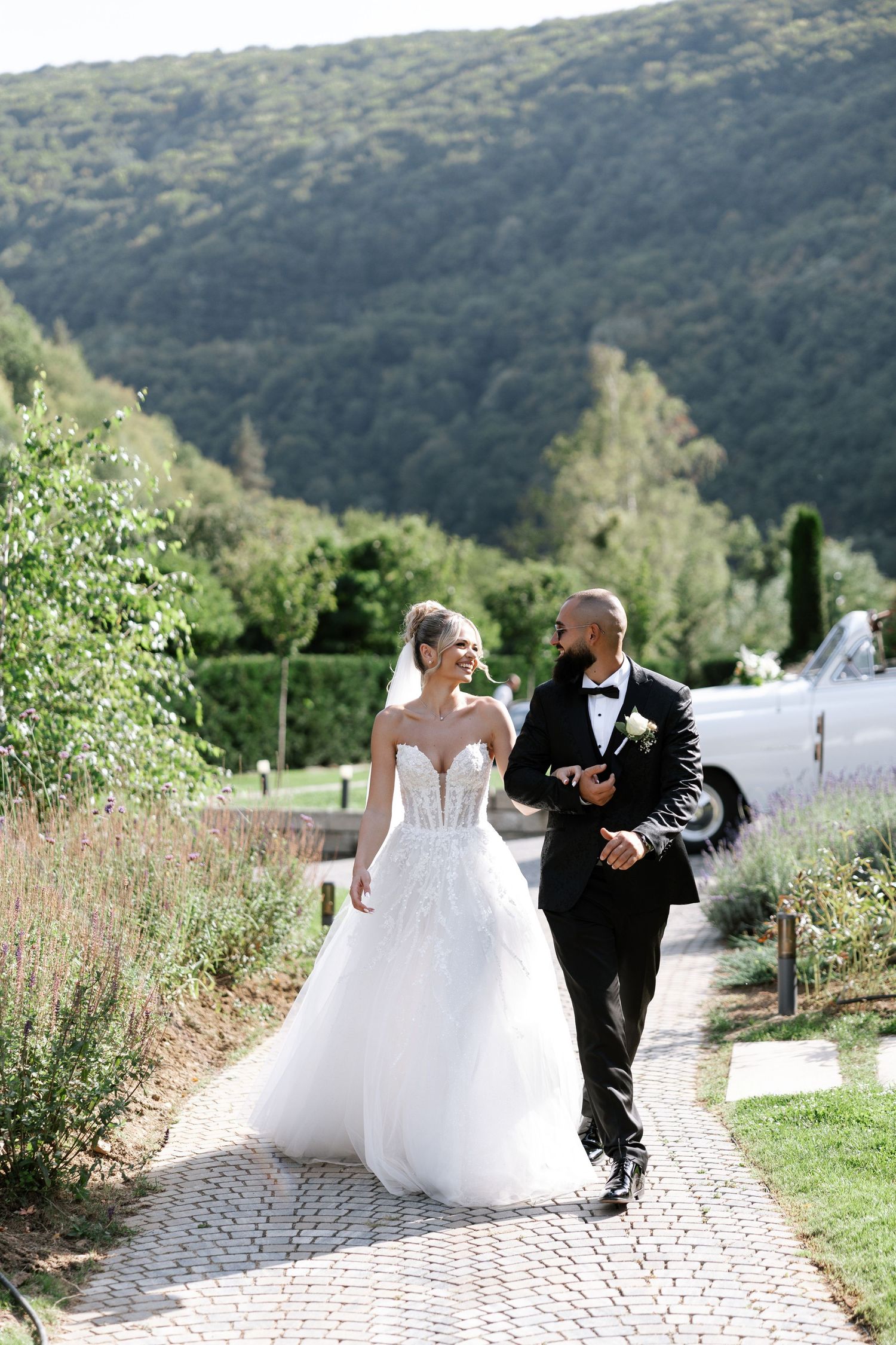 A couple walks along a garden path with lush green mountains in the background during their outdoor wedding celebration.