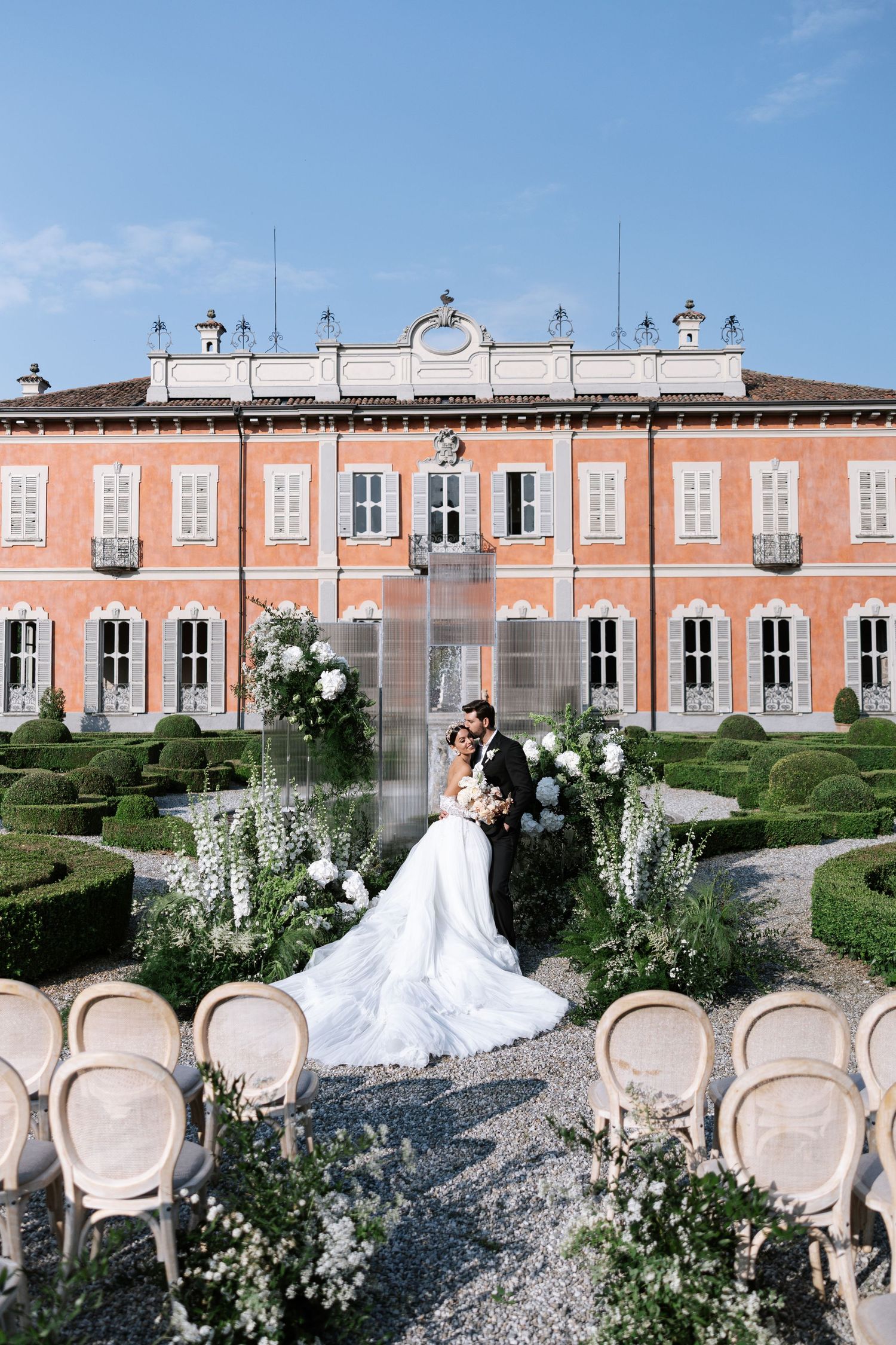 Elegant wedding ceremony setup in front of a pink Italian villa with manicured gardens and vintage chairs arranged outdoors.