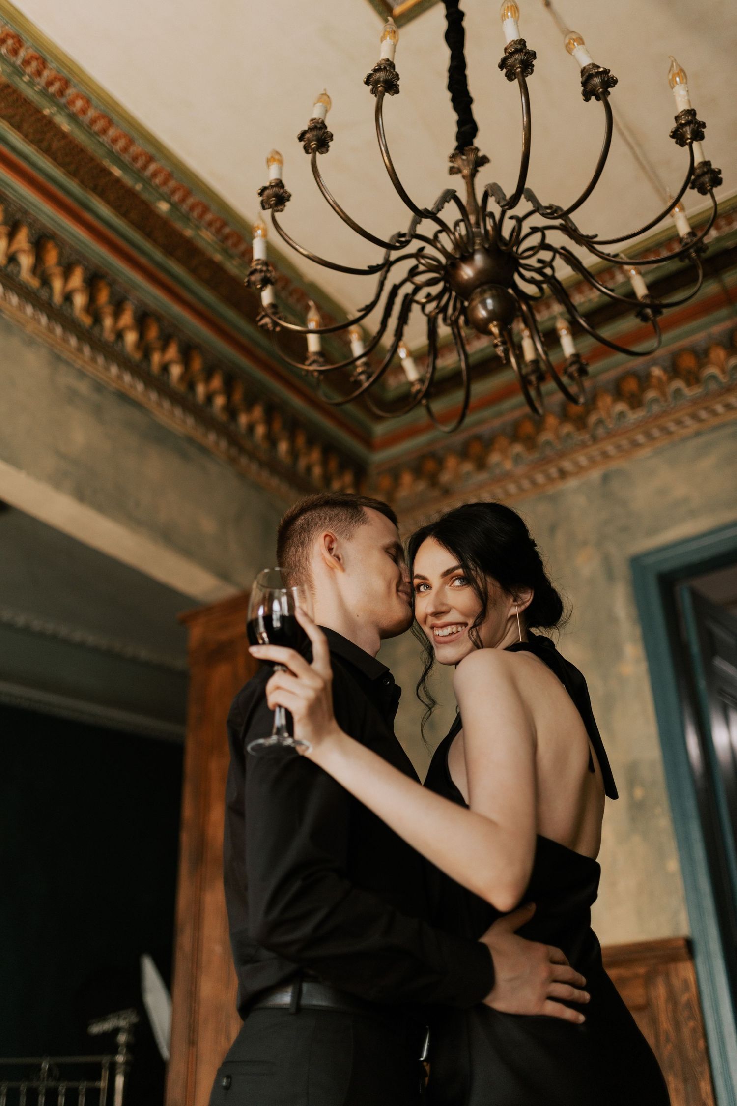 Couple in formal attire dance beneath ornate chandelier in vintage room.